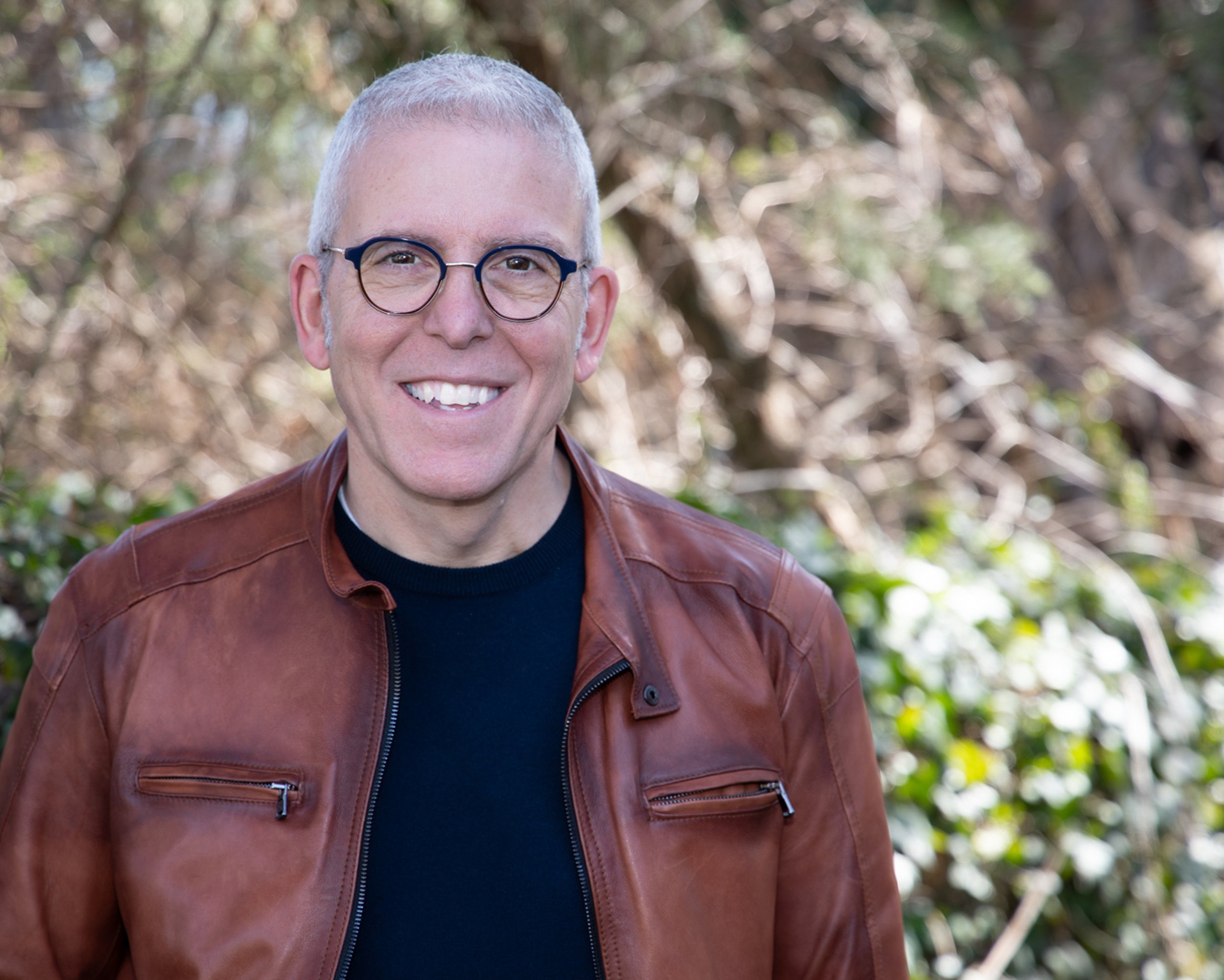 A man smiling outdoors with trees in the background, wearing glasses, a black shirt, and a brown leather jacket.