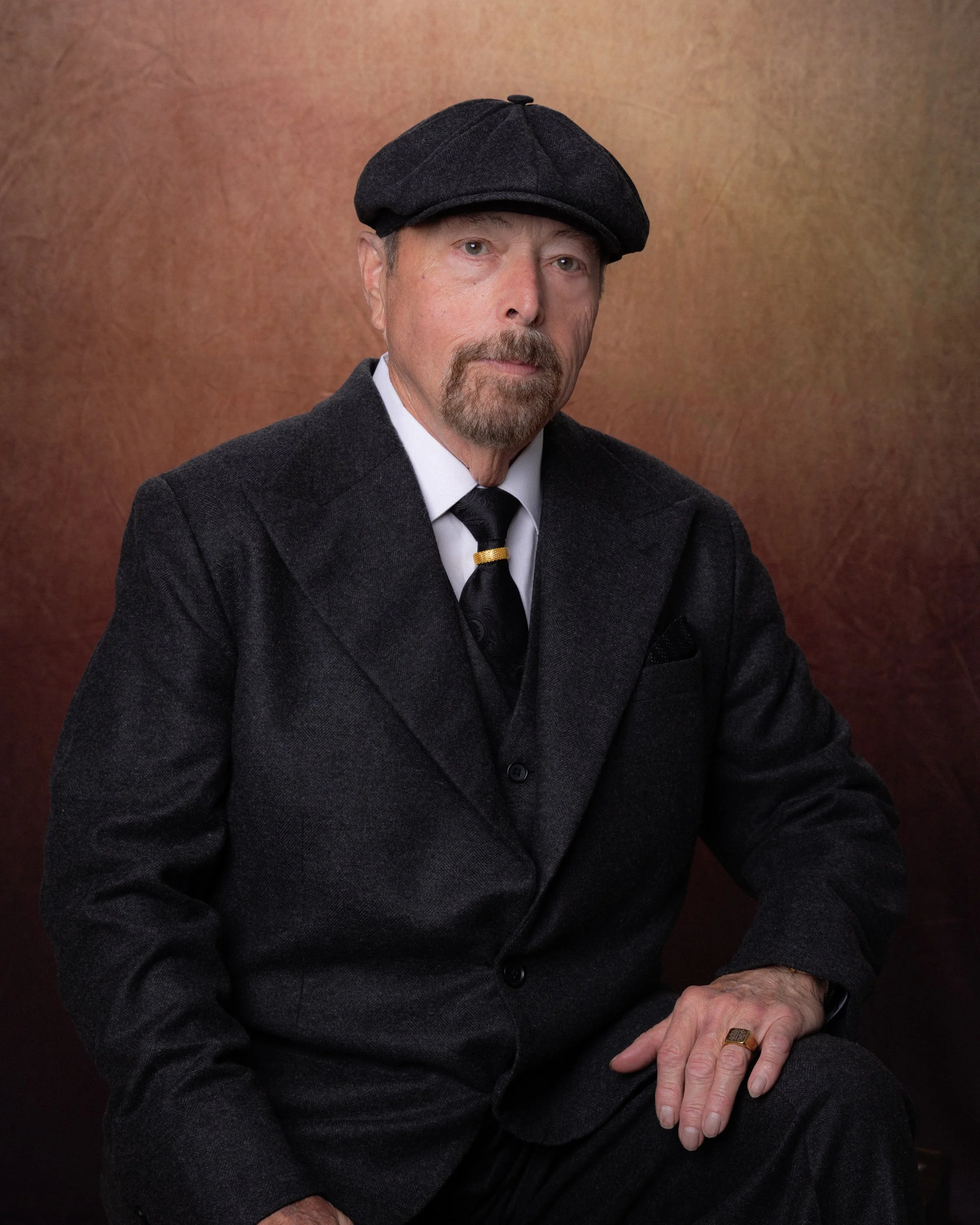 A portrait of an elderly man wearing a dark flat cap, a black coat, white shirt, and a black tie with a gold ring on his finger, sitting against a plain brownish background.