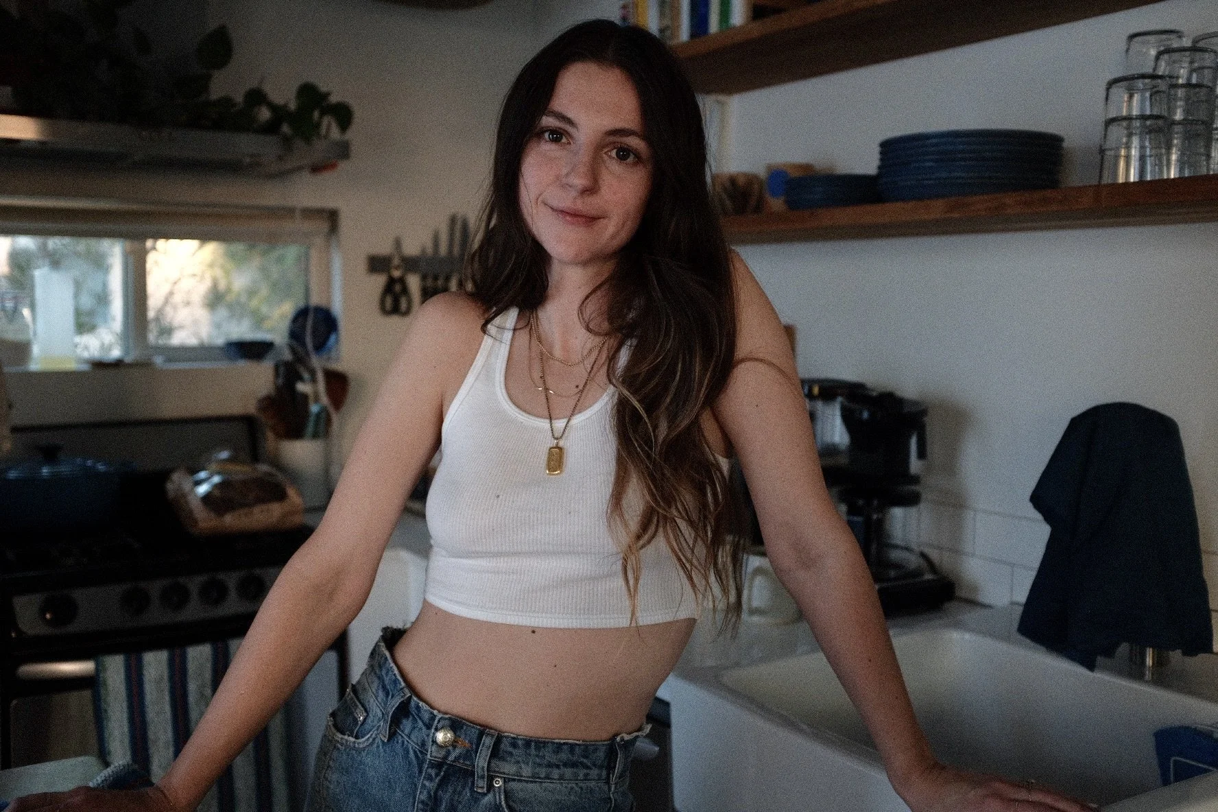 A young woman with long brown hair and freckles, wearing a white crop top and denim shorts, standing in a kitchen with a slight smile, with kitchen shelves and appliances in the background.
