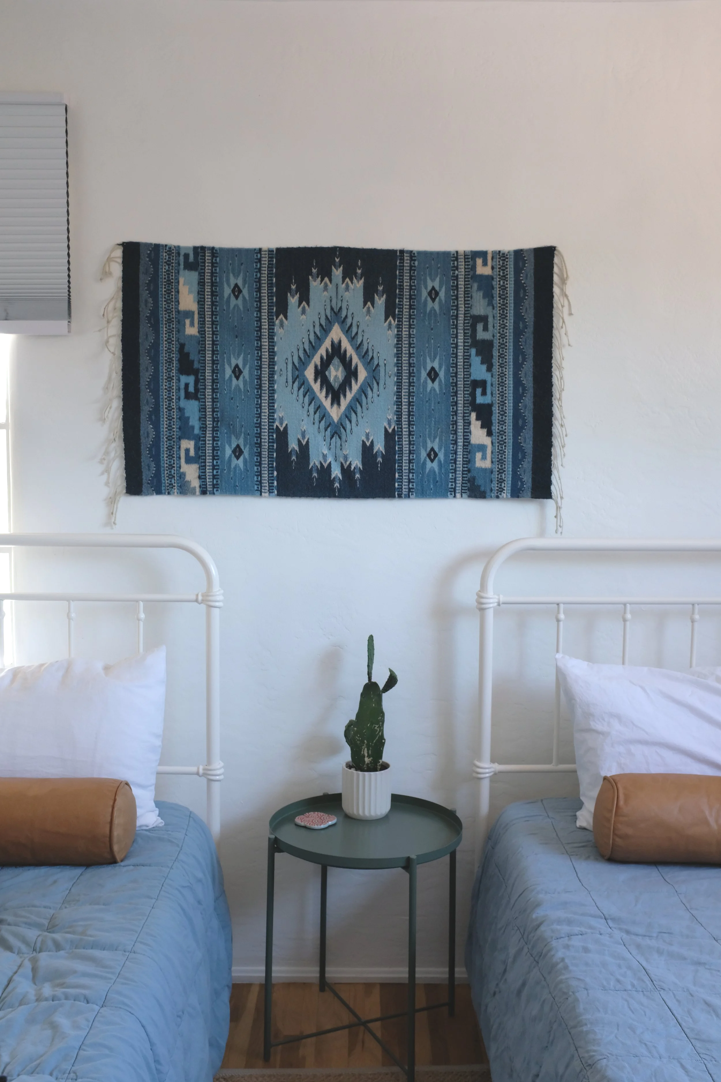 Two beds with light blue quilts and white pillows in a bedroom. Between the beds is a round table with a potted cactus and small decorative items. On the wall above is a blue patterned textile or wall hanging, and to the left is a window with closed white blinds.