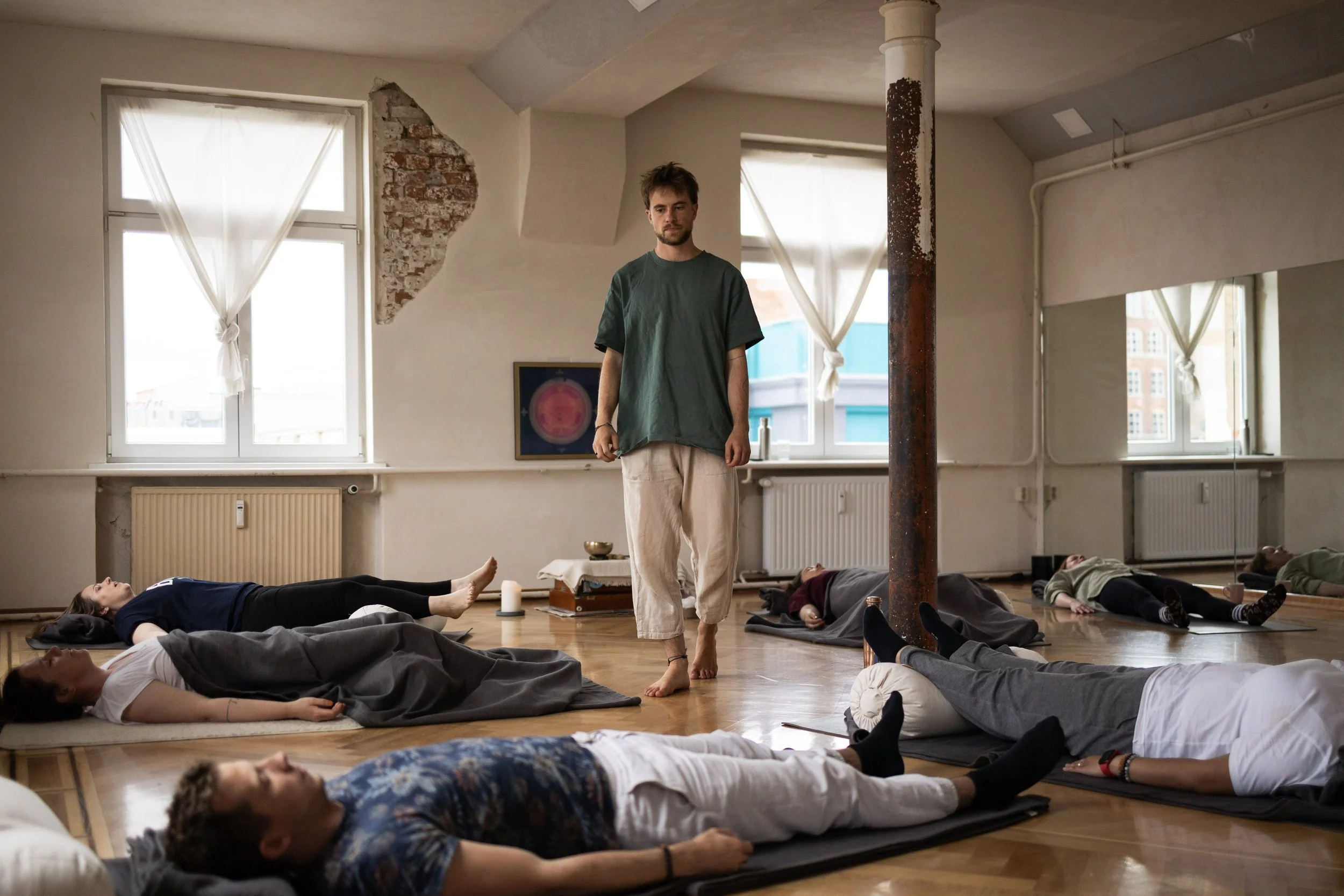 Ein junger Mann steht barfuß in der Mitte eines Raumes mit hohen Fenstern und einer alten Mauer, in der Yoga oder Meditation praktiziert wird, während mehrere Menschen auf Yogamatten liegen, in einer entspannten Pose.