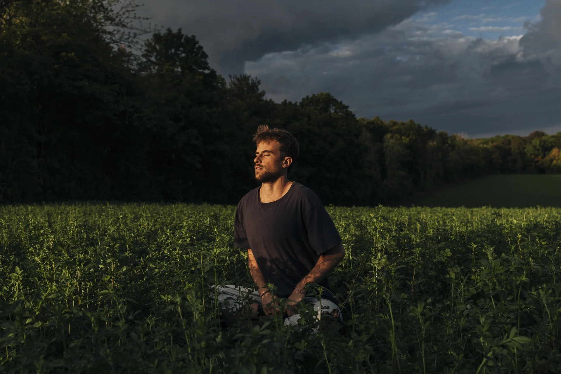 Junge Person, die im Feld bei Sonnenuntergang oder Sonnenaufgang Atemübungen praktiziert, umgeben von grünen Pflanzen, dunklen Wolken am Himmel.
