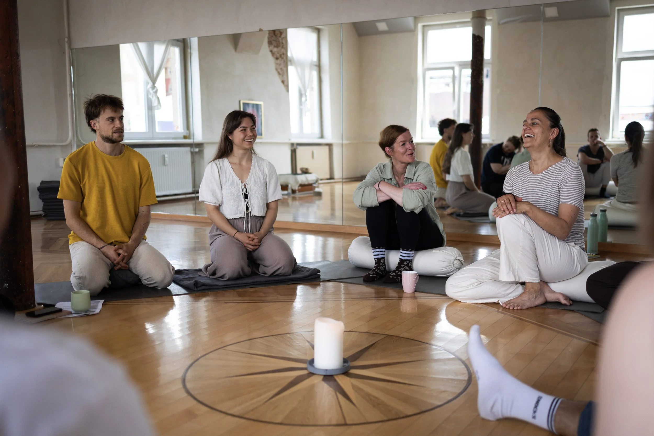 Gruppe von Menschen in einem Raum, die auf einem Boden sitzen und lachen, möglicherweise bei einer gemeinsamen Aktivität oder einem Workshop.