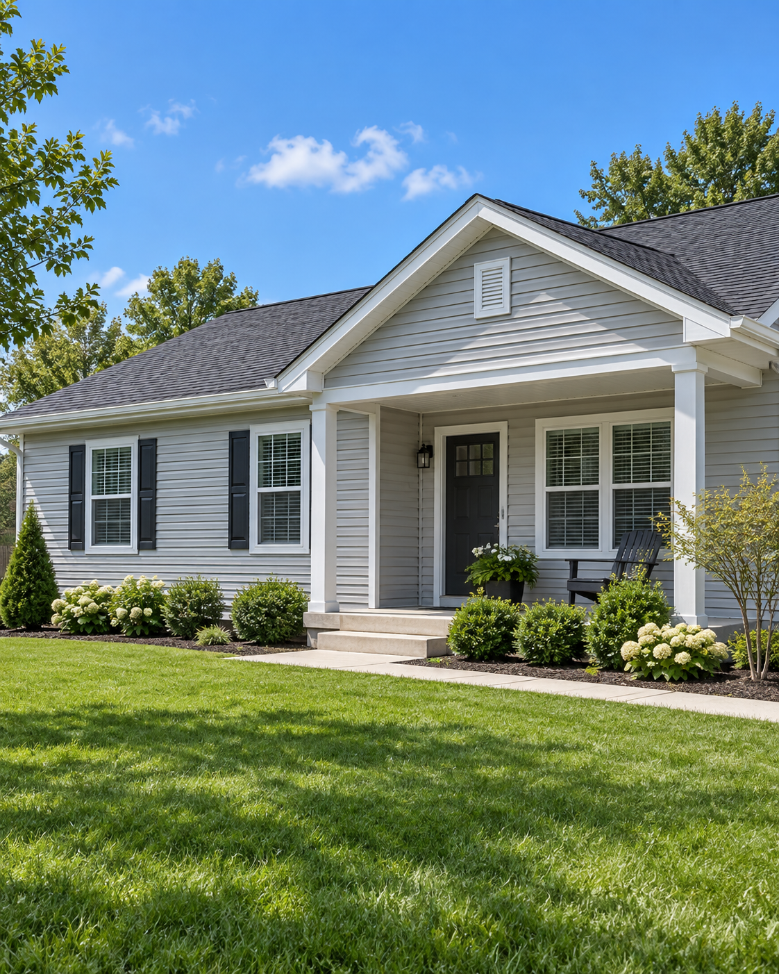 Front view of a single-story house with light gray siding, black shutters, and a dark gray roof. The house has a covered porch, white trim, a black front door, and landscaped bushes and flowers in the yard. Bright green grass and a blue sky with a few clouds are visible.