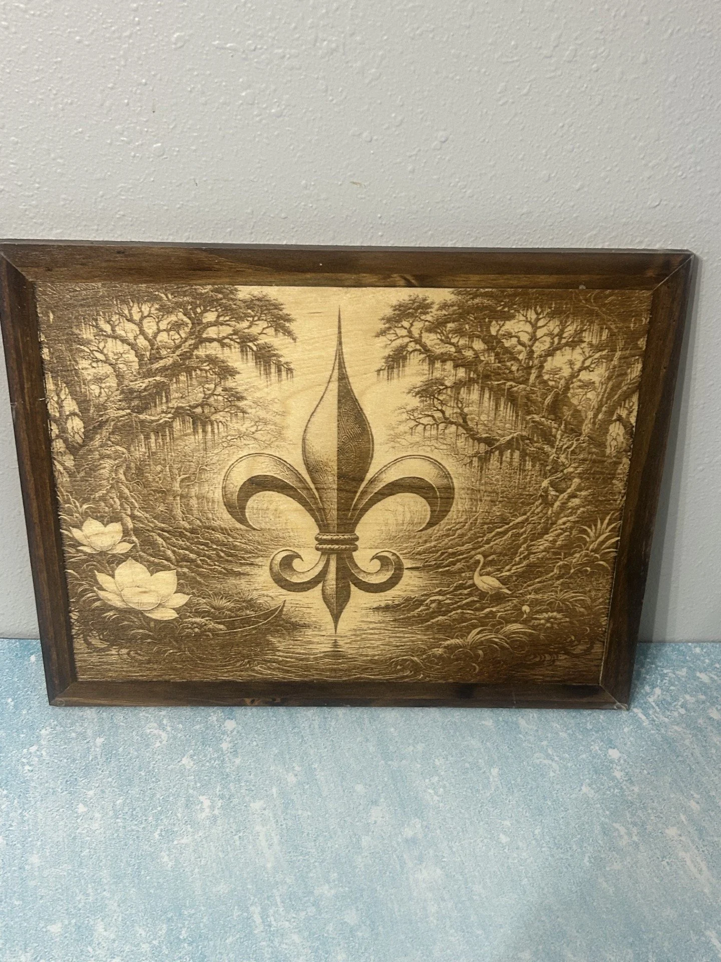 Wooden wall art with a fleur-de-lis symbol in the center, surrounded by trees, ducks, and water lilies, framed with a dark wooden border.