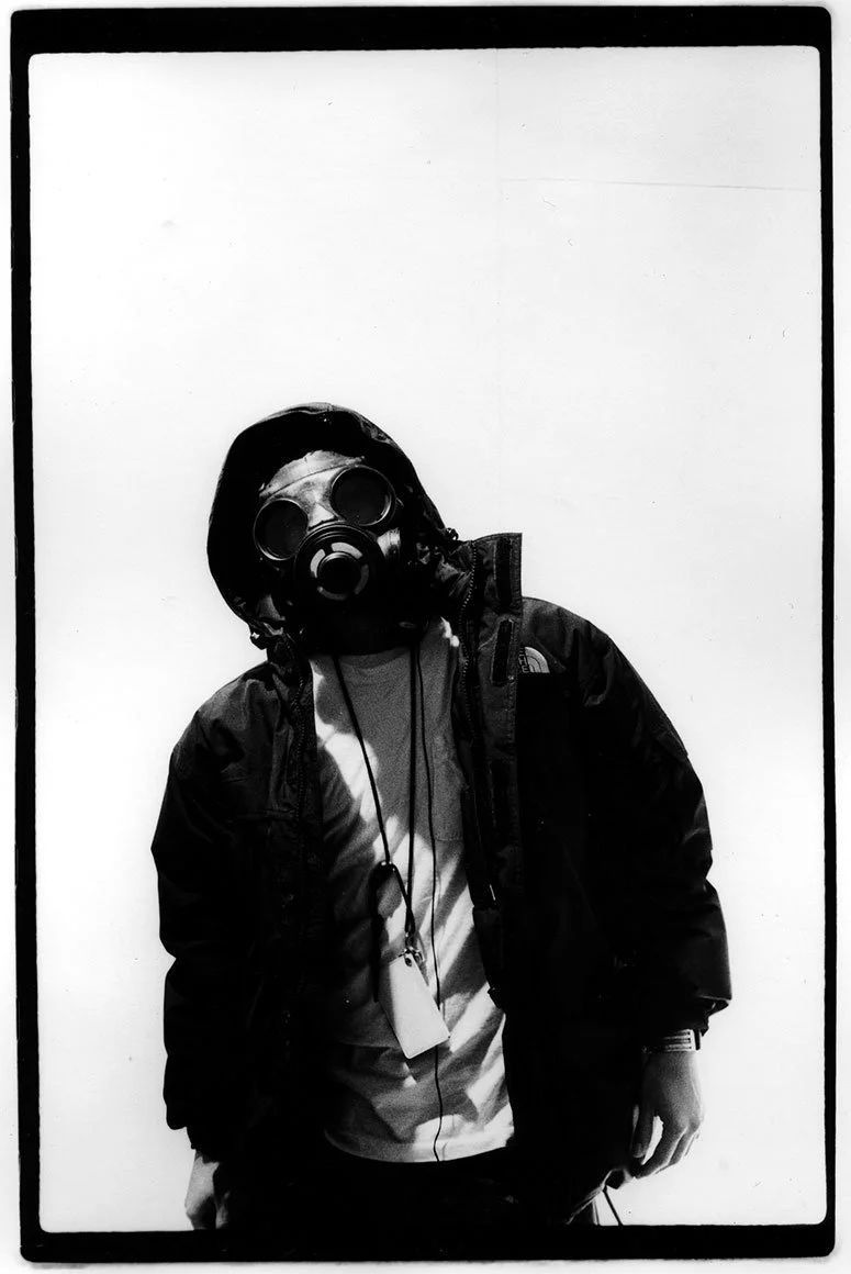 A person wearing a gas mask, hoodie, and jacket standing against a plain white background.