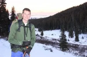 A man standing outdoors in a snowy, mountainous landscape with pine trees, likely on a winter hike or exploration.
