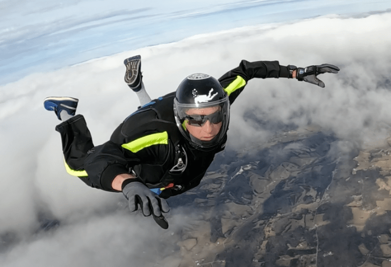 Skydiver in black jumpsuit with neon yellow stripes, wearing a helmet and goggles, free-falling above a cloudy landscape.