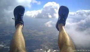A person skydiving with legs and blue shoes visible, descending with clouds and a cityscape below.