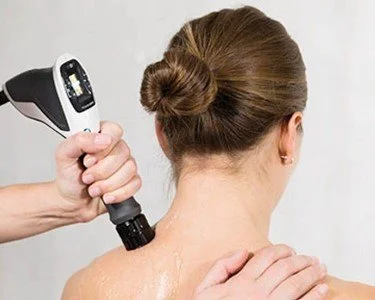 A person receiving a back massage while another person uses a handheld device on their shoulder in a spa or massage setting.