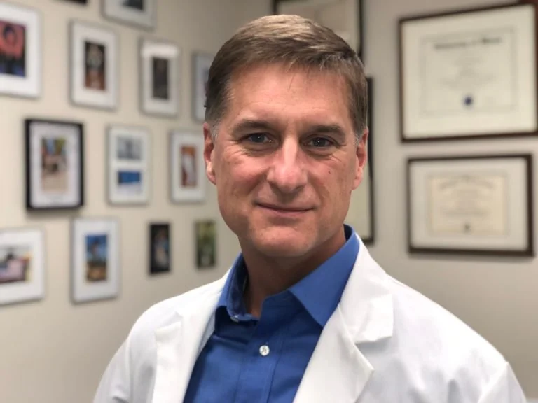 A male doctor in a white coat standing in an office or clinic with framed certificates and photographs on the wall behind him.