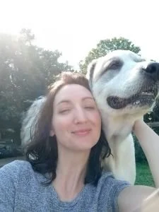 Young woman smiling and hugging a large white dog outdoors on a sunny day.