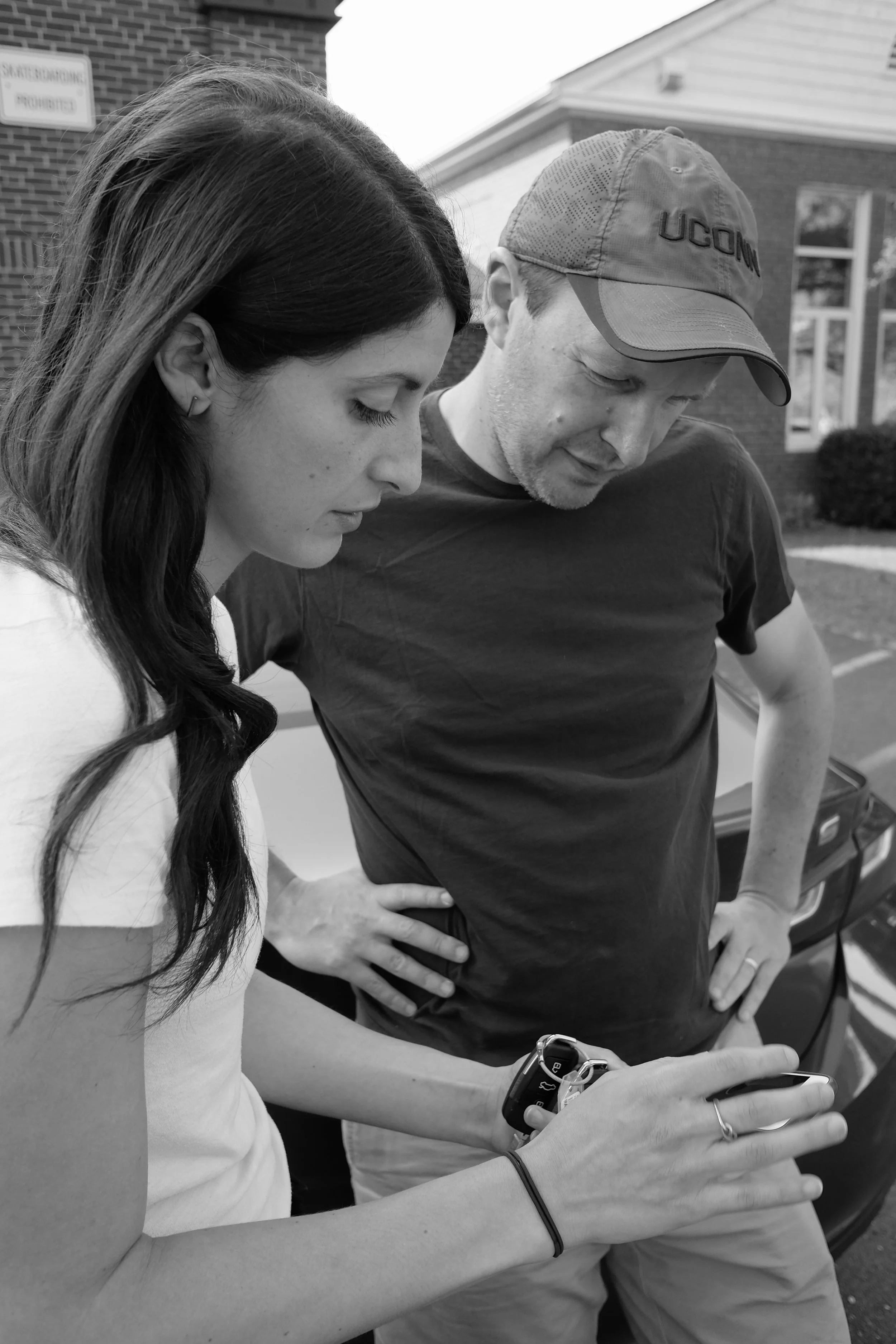 A young woman and a middle-aged man stand outside, looking at a smartphone together. The woman has long, dark hair, and the man wears a UConn cap and a t-shirt. They are focused on the phone, which the woman is holding.