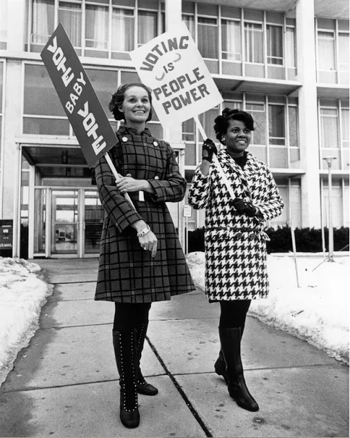 Two women holding signs advocating for voting rights and women’s power, standing on a sidewalk in front of a modern building, with snow on the ground.