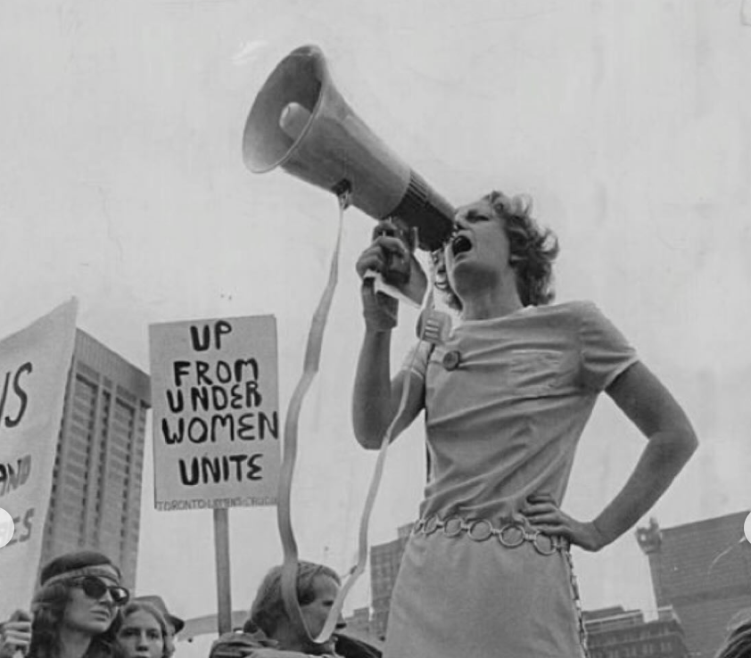 A woman speaking into a megaphone at a protest or rally, surrounded by other protesters holding signs, including one that says 'UP FROM UNDER WOMEN UNITE,' in an urban setting.