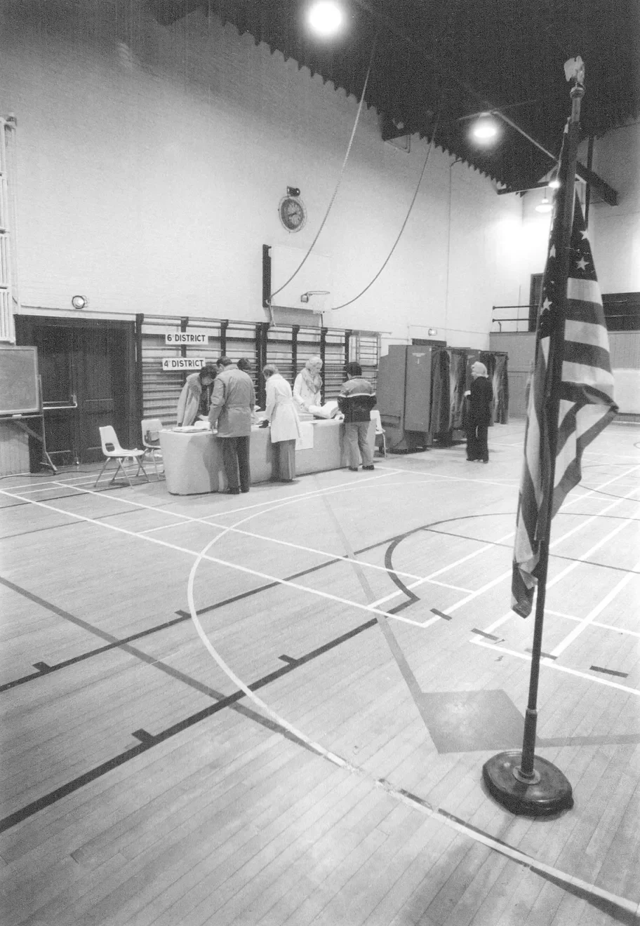 A vintage black and white photo of a polling station inside a gymnasium, with people voting at a table marked '6th District' and '4th District,' and an American flag in the foreground.