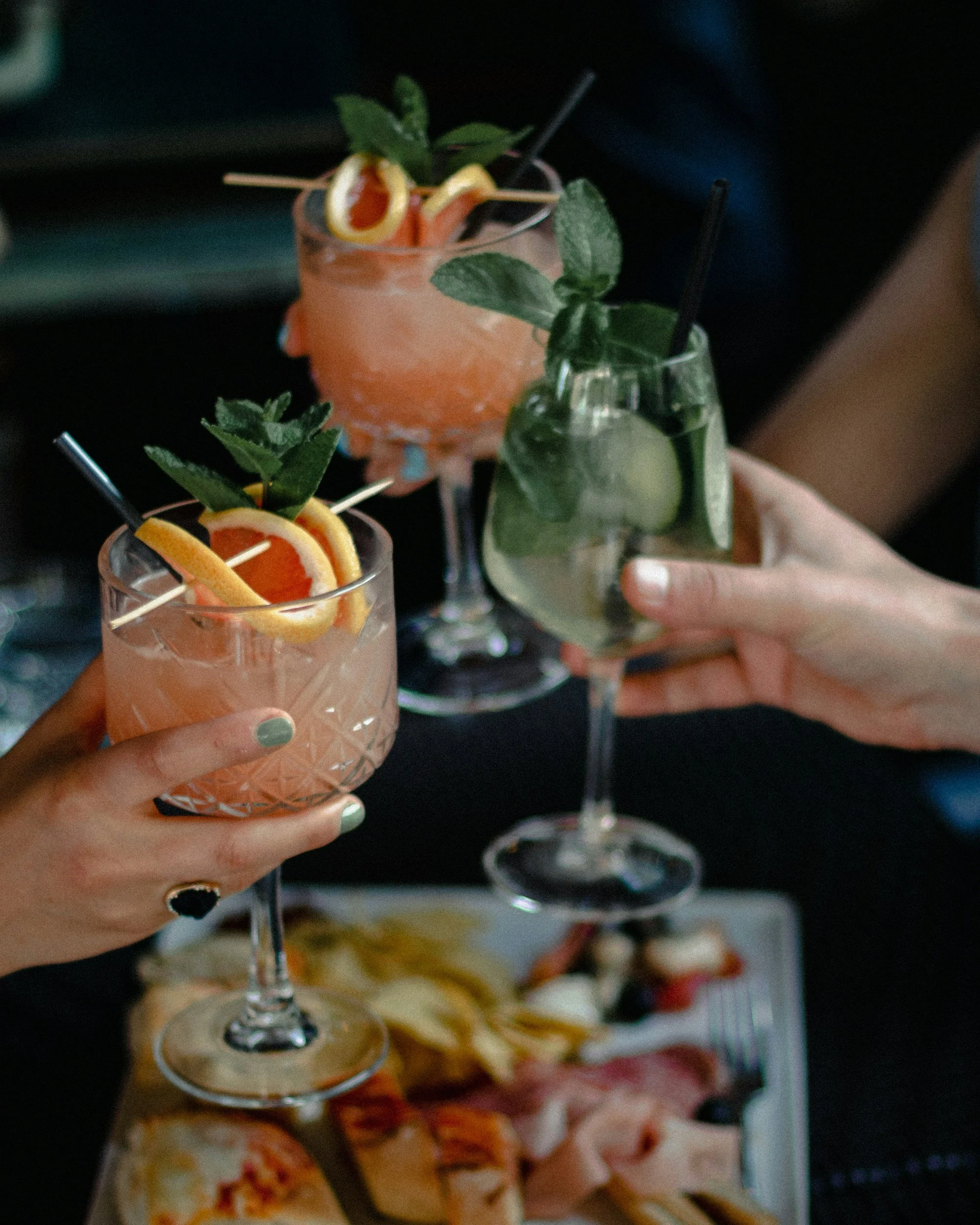 Close-up of a hand holding a pink cocktail glass garnished with lemon slices, herbs, and a skewer, with other drinks and appetizers in the background.