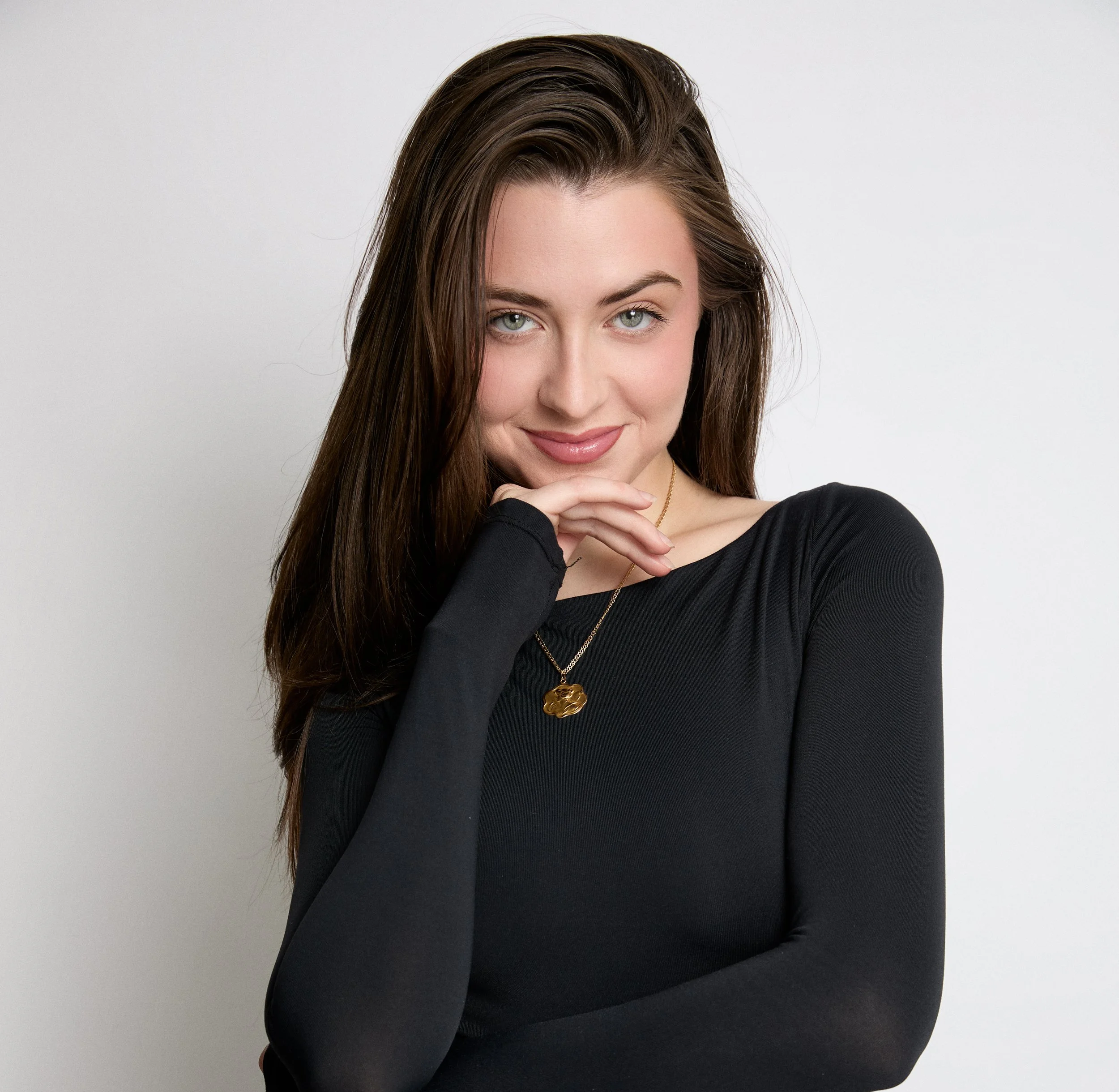 A woman with long brown hair and light green eyes is smiling softly. She is wearing a black top and a gold necklace with a pendant. Her right hand is touching her chin, and the background is plain white.