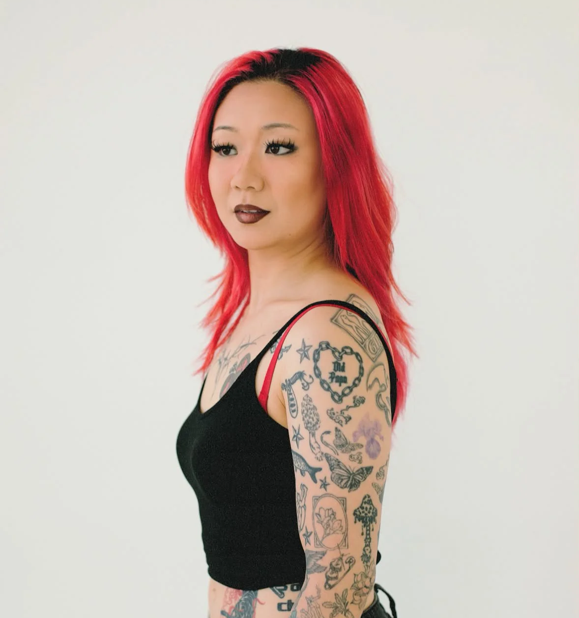 A woman with red hair and tattoos on her arm, wearing a black tank top with red straps, standing against a plain white background.