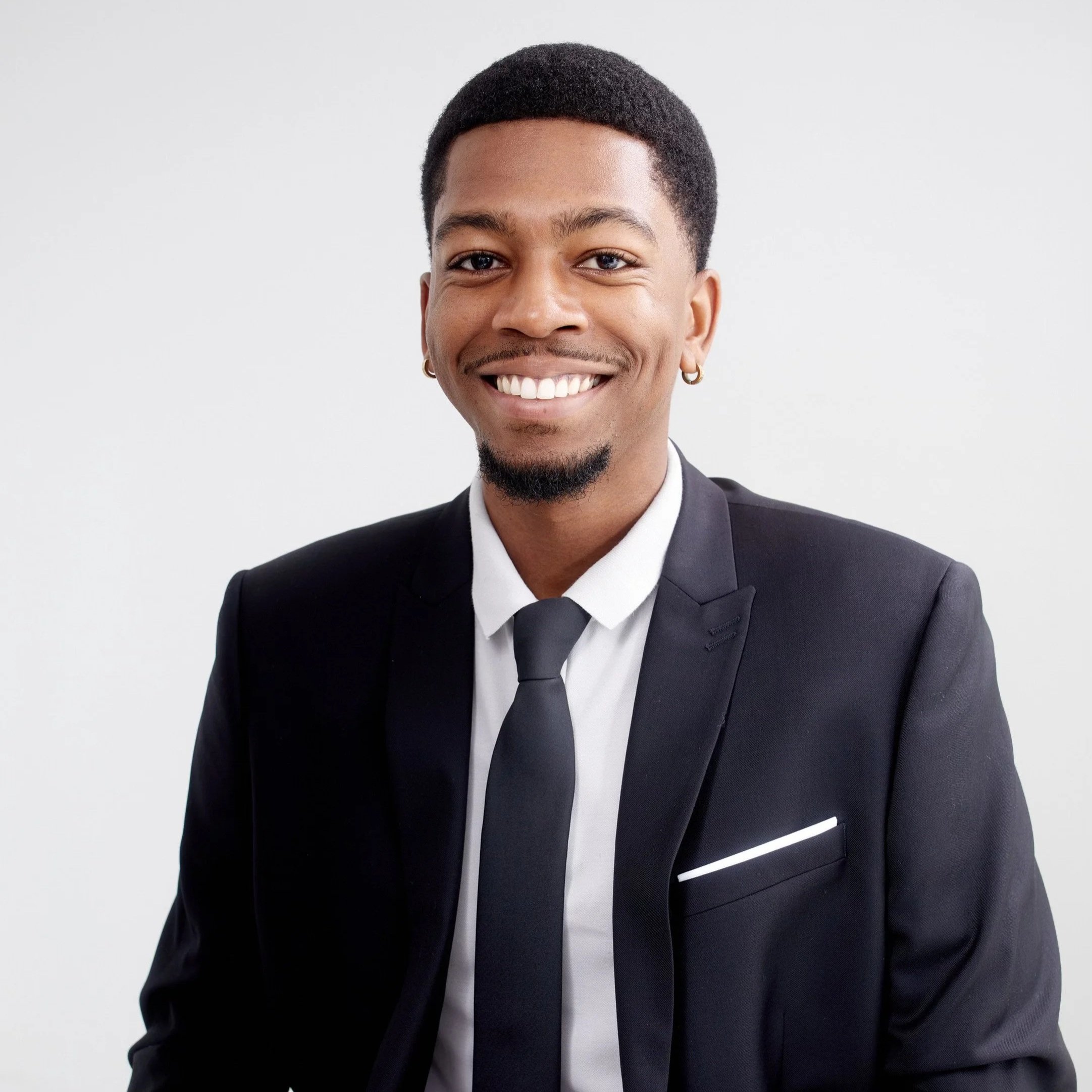 Close-up of a smiling man with short black hair, earrings, and a goatee, wearing a black tuxedo with a white shirt and black bow tie, against a plain light background.