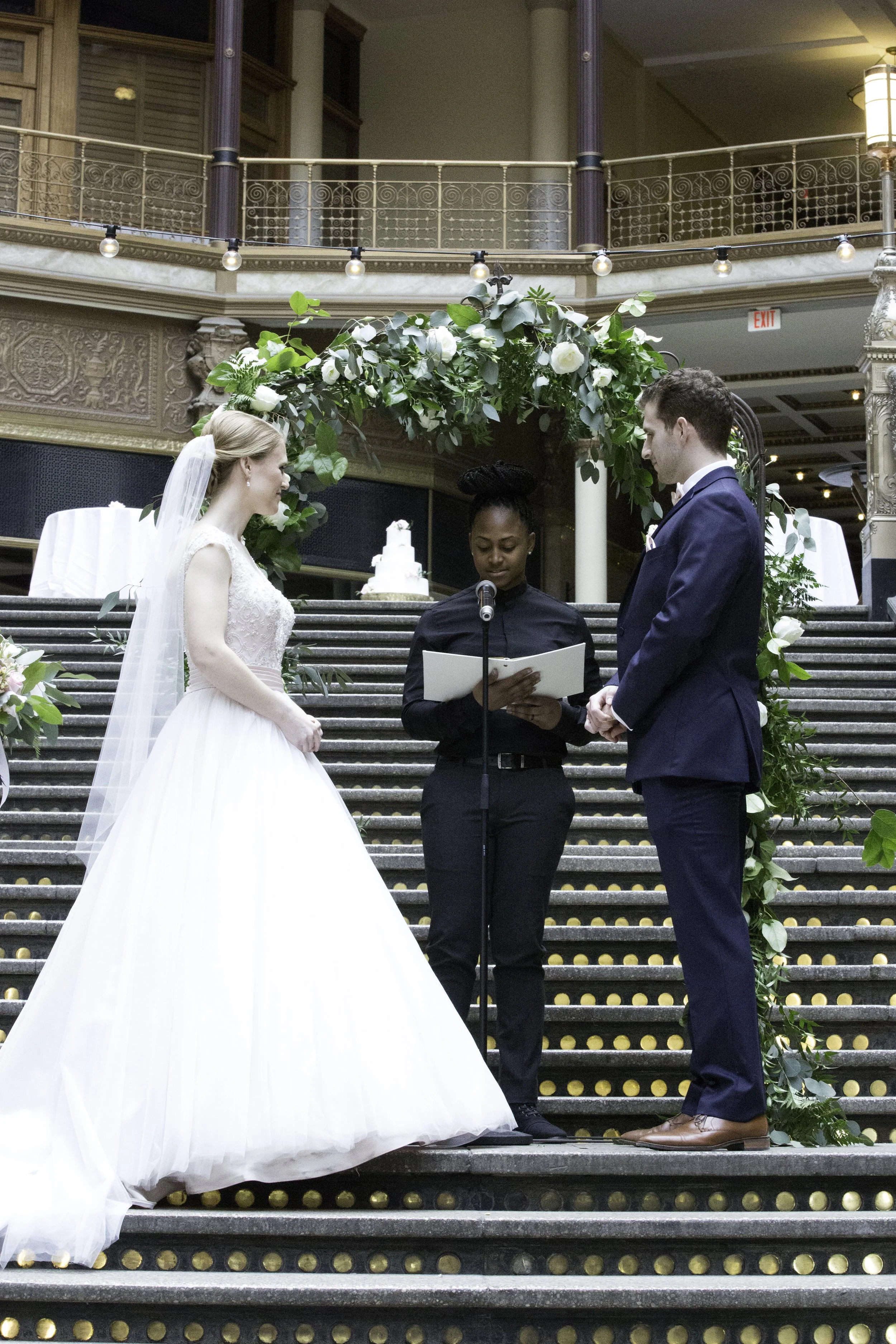 A bride and groom stand on a decorated staircase during a wedding ceremony, with an officiant reading from a book. The bride wears a white wedding gown with lace detail and a veil, and the groom wears a navy blue suit. There is a large floral arrangement with white flowers and greenery behind them, and a wedding cake is visible in the background.