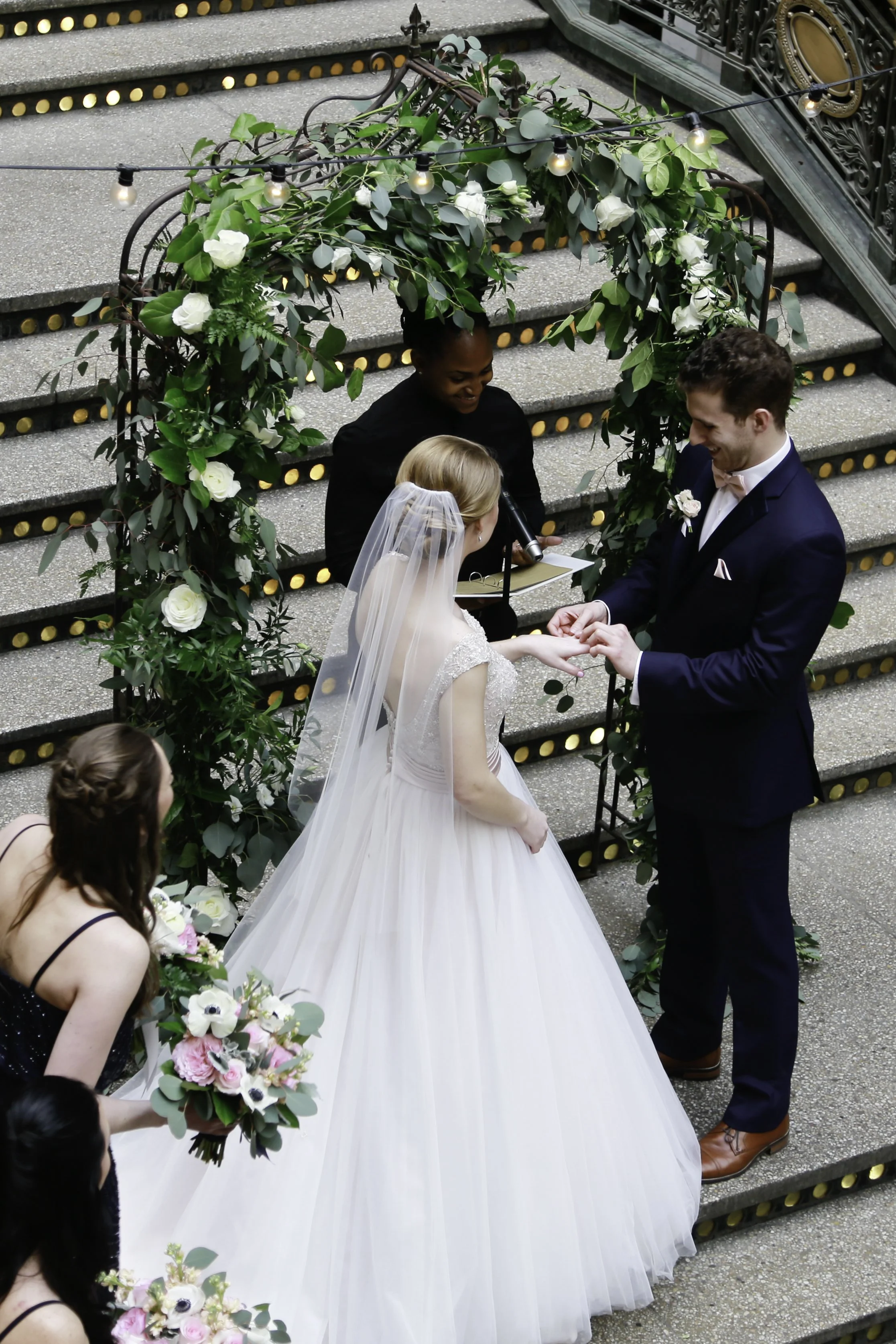 A bride and groom exchanging rings during their wedding ceremony on a staircase decorated with greenery and white flowers, with bridesmaids holding bouquets nearby and an officiant officiating.
