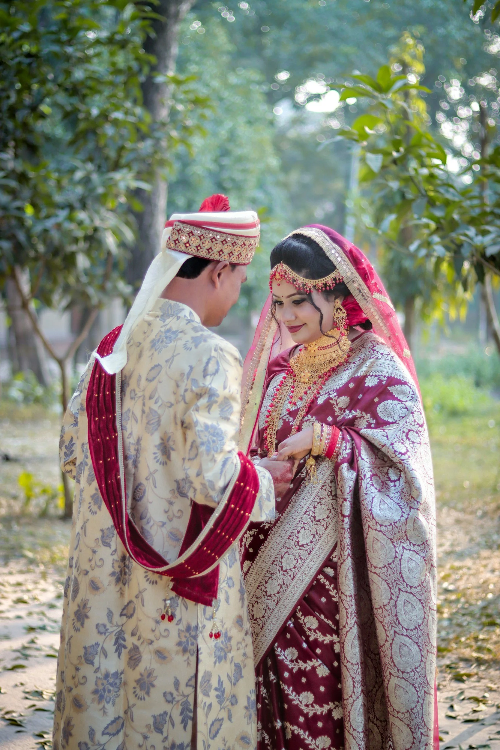 A bride and groom in traditional Indian wedding attire exchanging rings outdoors, surrounded by trees and greenery.