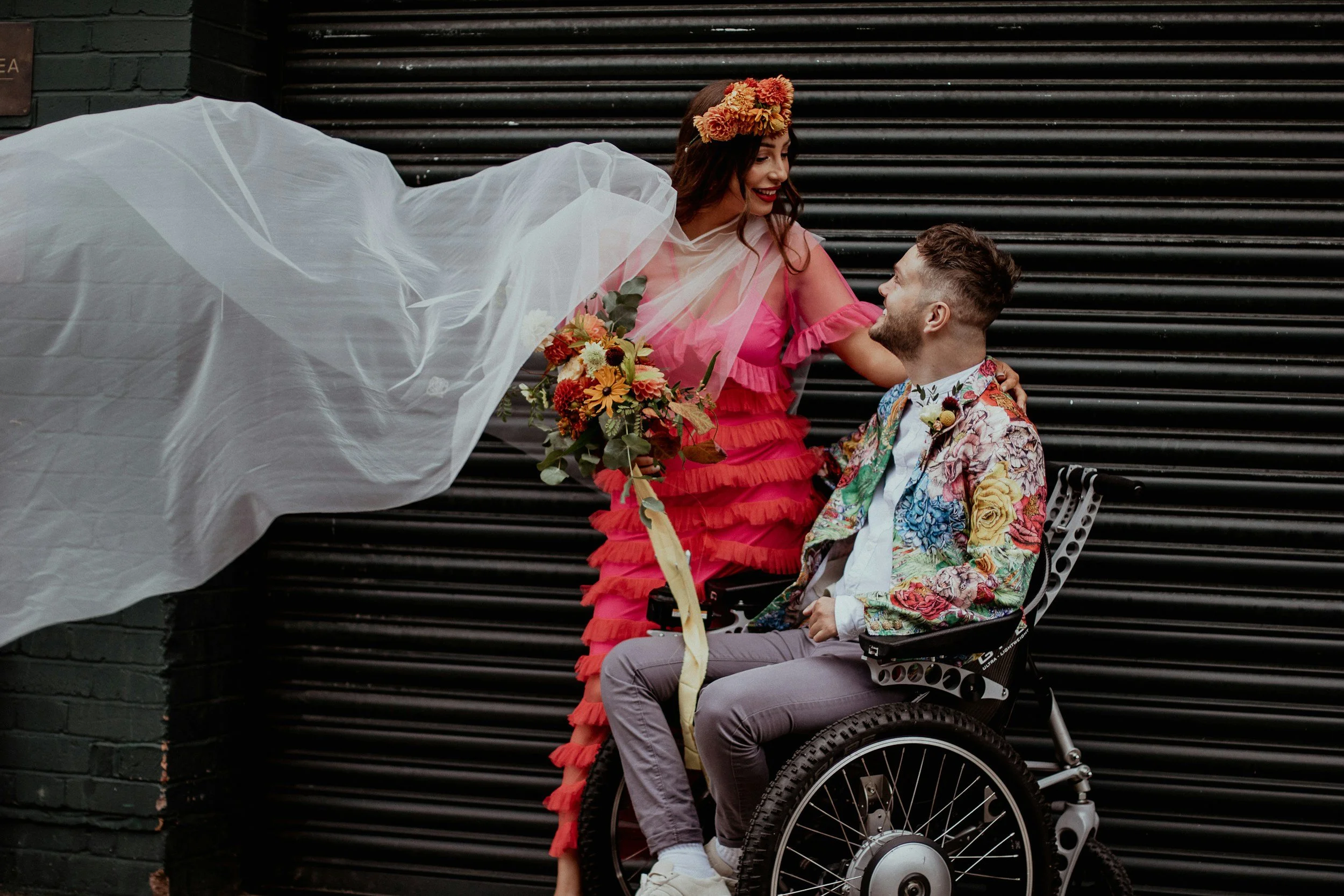 A woman in a floral dress with a flower crown smiling at a man in a wheelchair wearing a colorful floral blazer, outdoors against a black metal shutter.