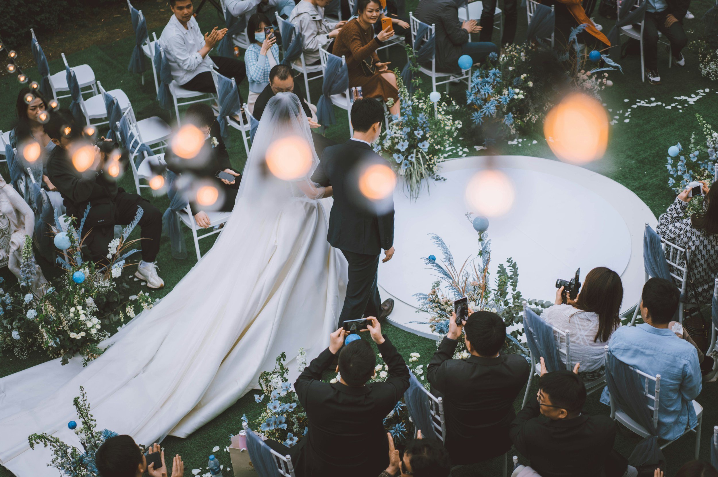 A wedding ceremony outdoors with a bride and groom standing under a floral arch, surrounded by seated guests taking photos and clapping.