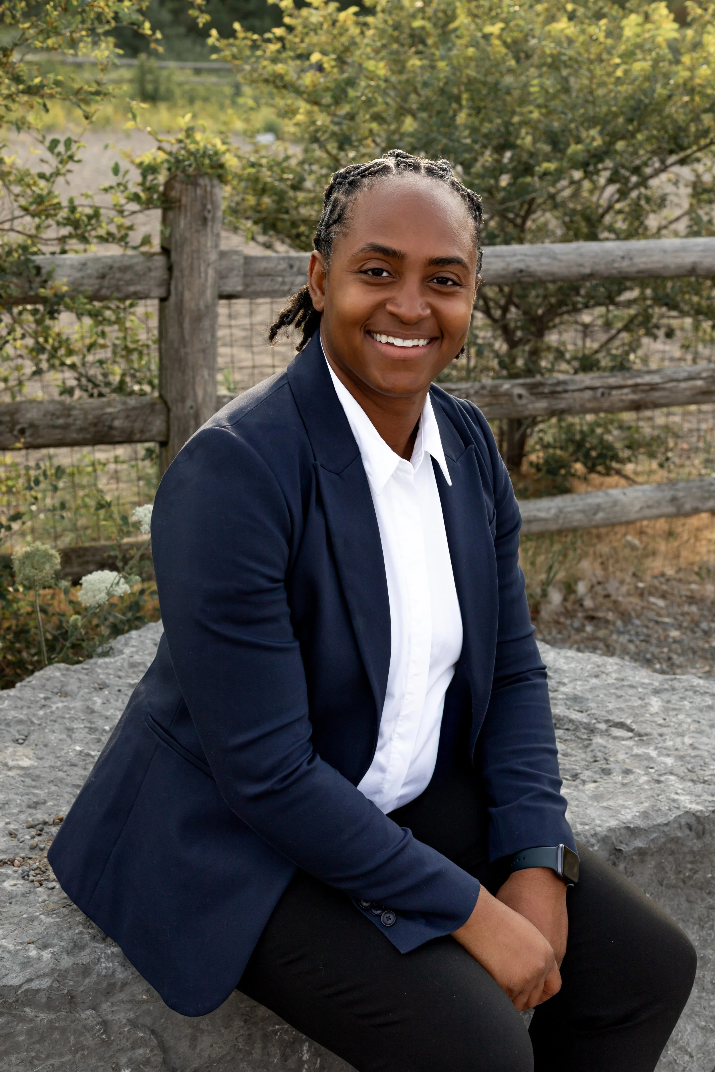 A woman with short braided hair, wearing a navy blazer and white shirt, sitting outdoors on a rock with a wooden fence and trees in the background, smiling at the camera.
