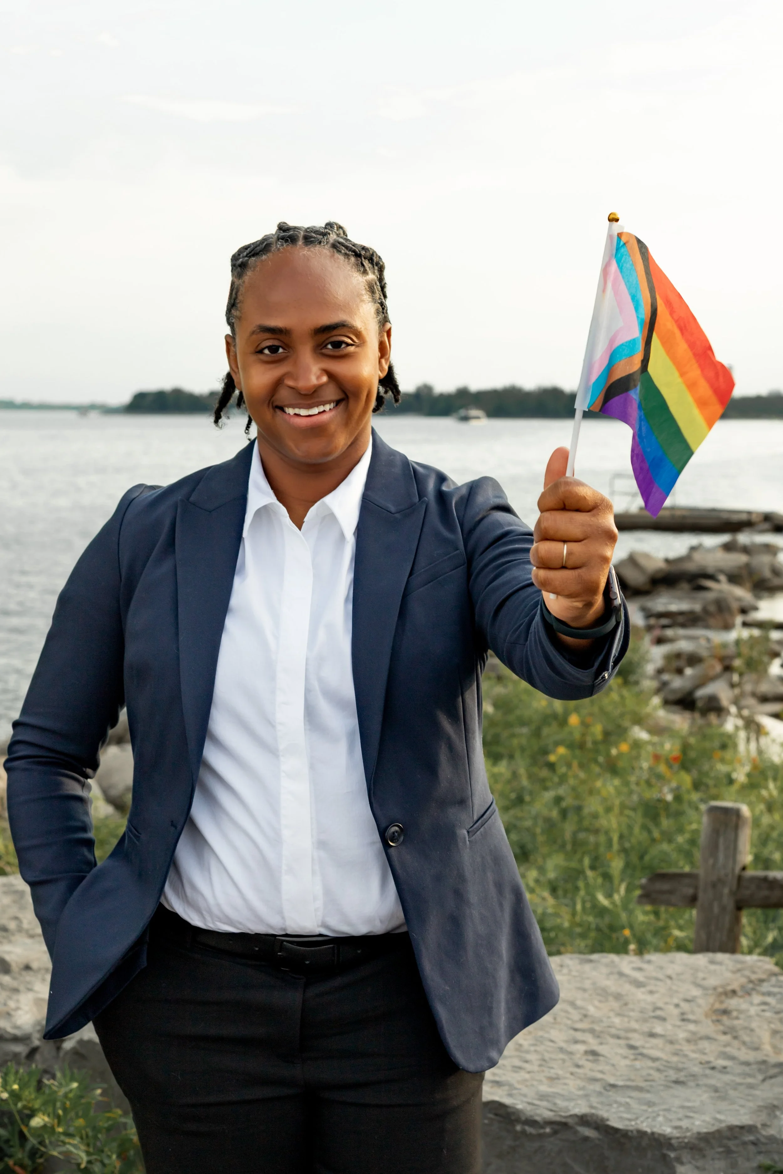 Woman in business attire holding a rainbow pride flag by a waterfront with clouds and trees in the background.