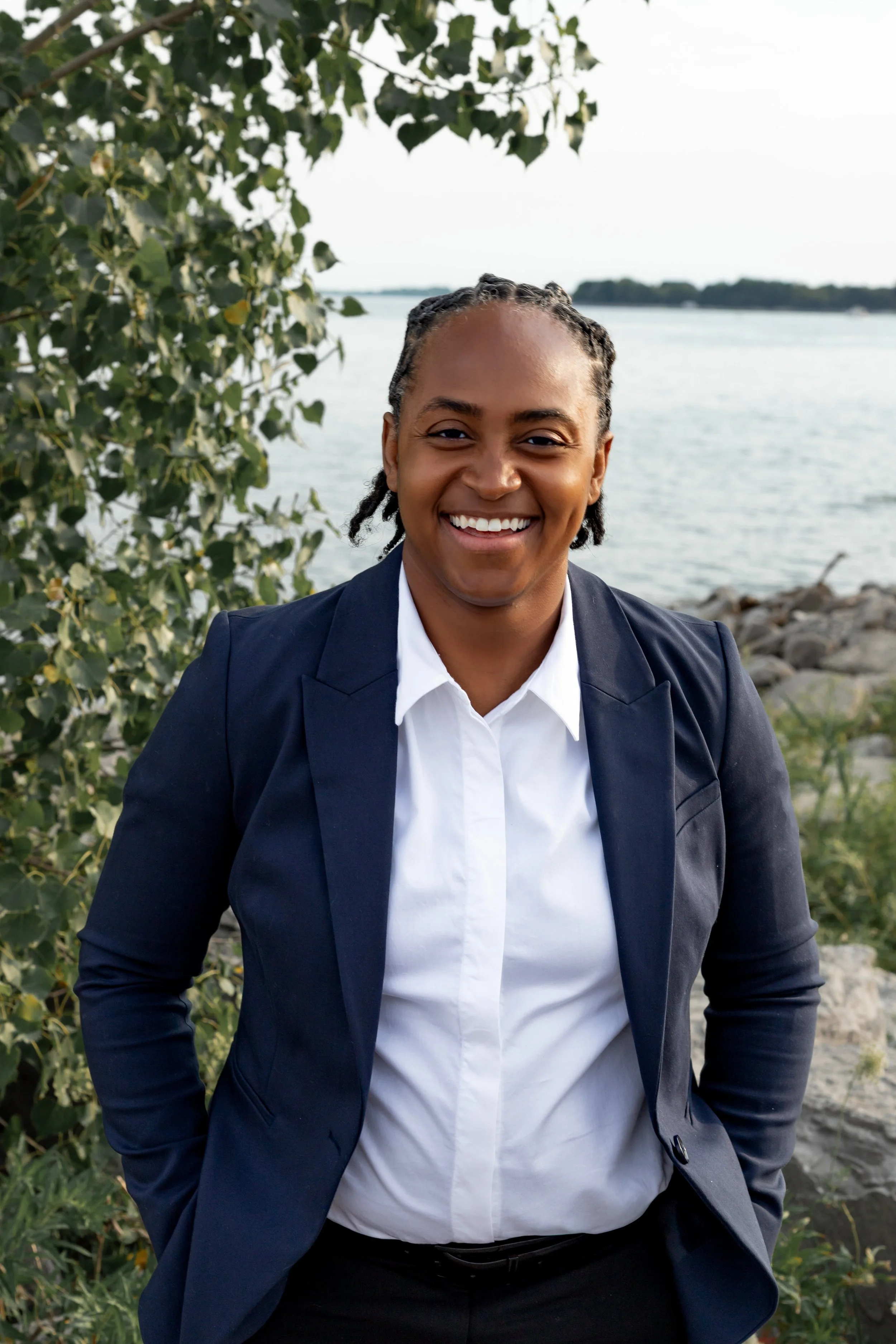 A smiling woman in a navy blue blazer and white shirt standing outdoors near a body of water with trees and rocks in the background.