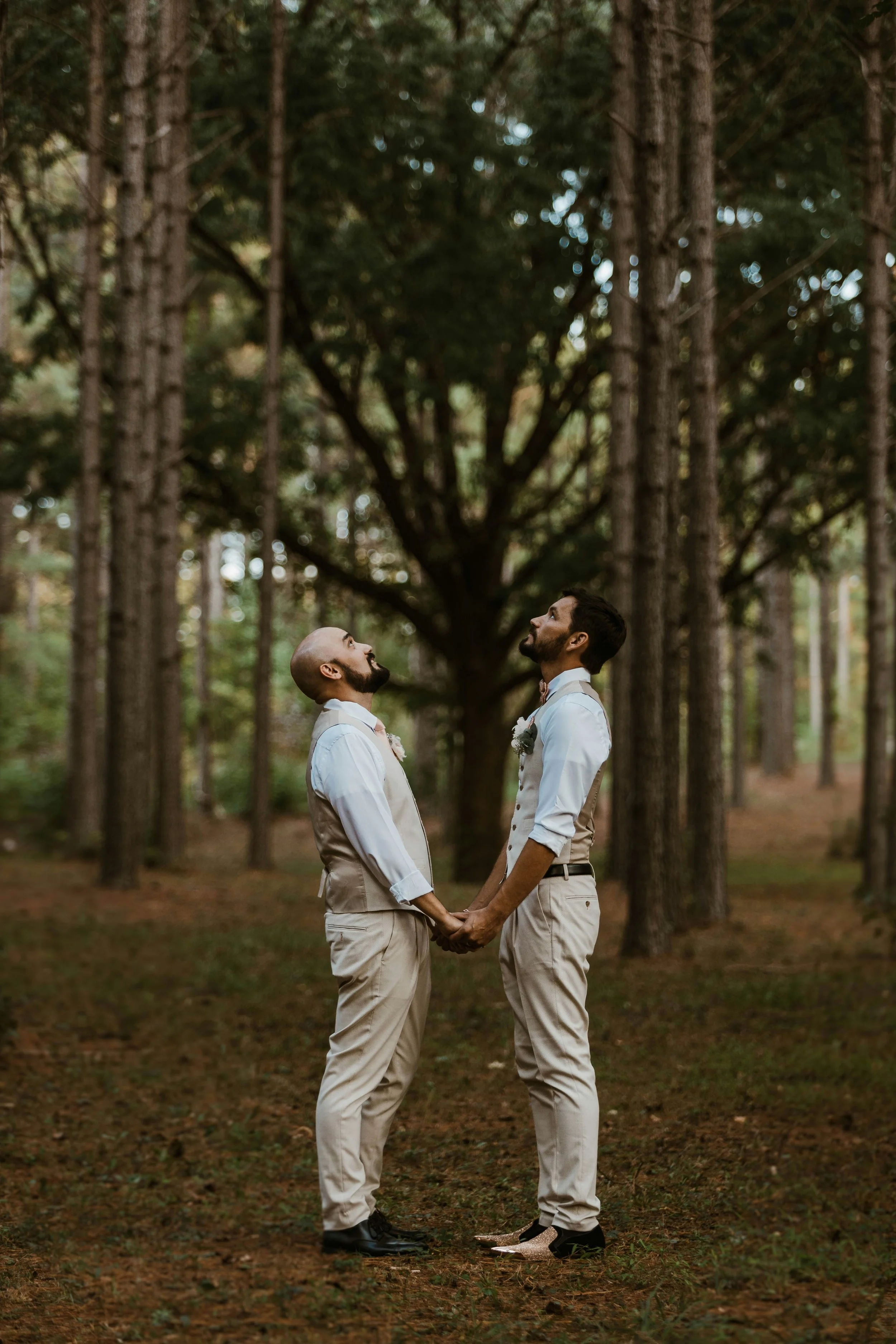 Two men in formal attire, holding hands and gazing into each other's eyes, standing in a forest with tall trees and green foliage.