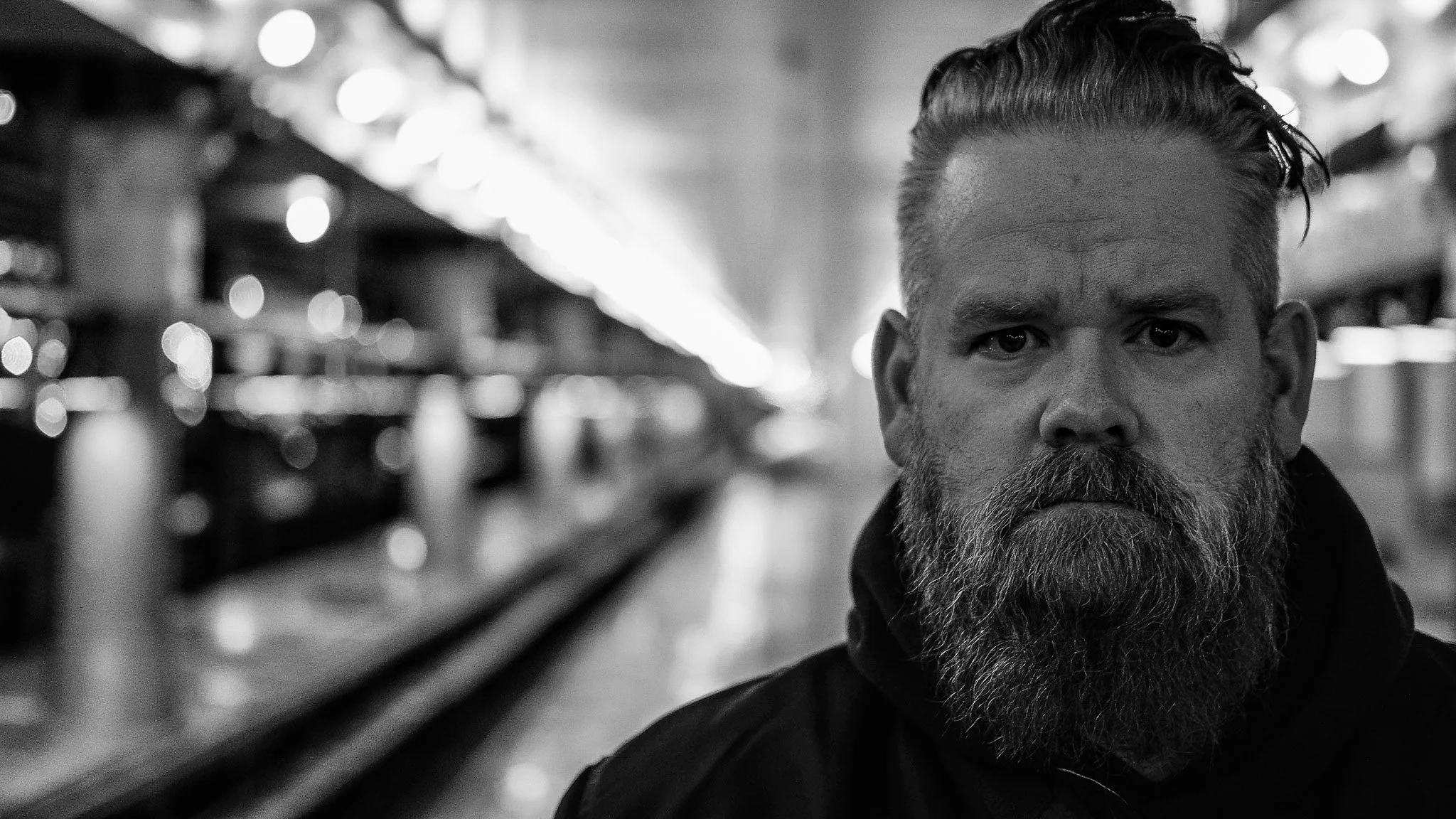 Black and white photo of a bearded man with styled hair in a dimly lit, blurred interior setting.