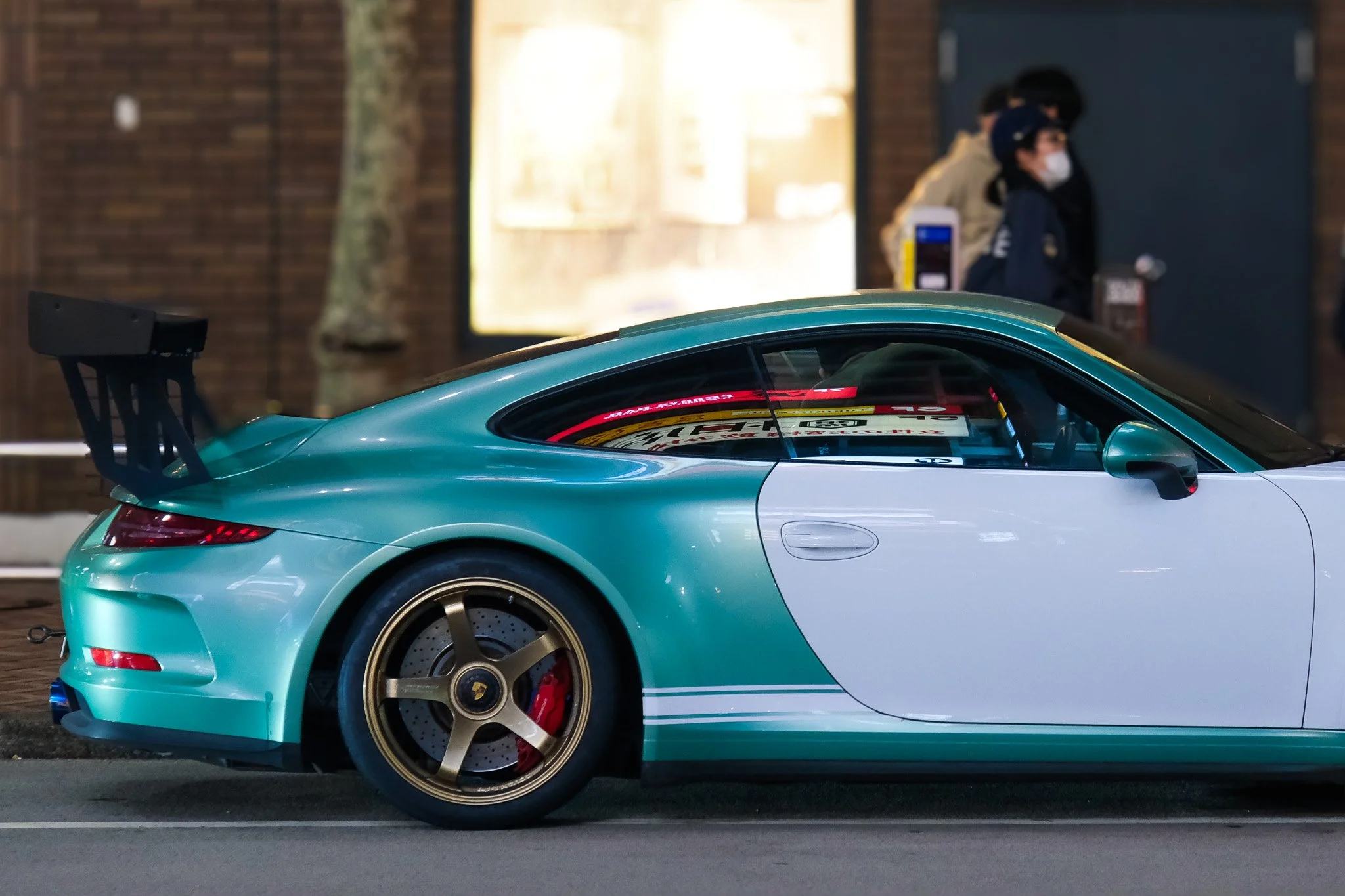 Close-up of a turquoise Porsche sports car with a large rear wing, gold wheels, and red brake calipers parked on a city street near Shibuya Crossing in Tokyo Japan. In the background, two police officers wearing masks and helmets write a ticket. 