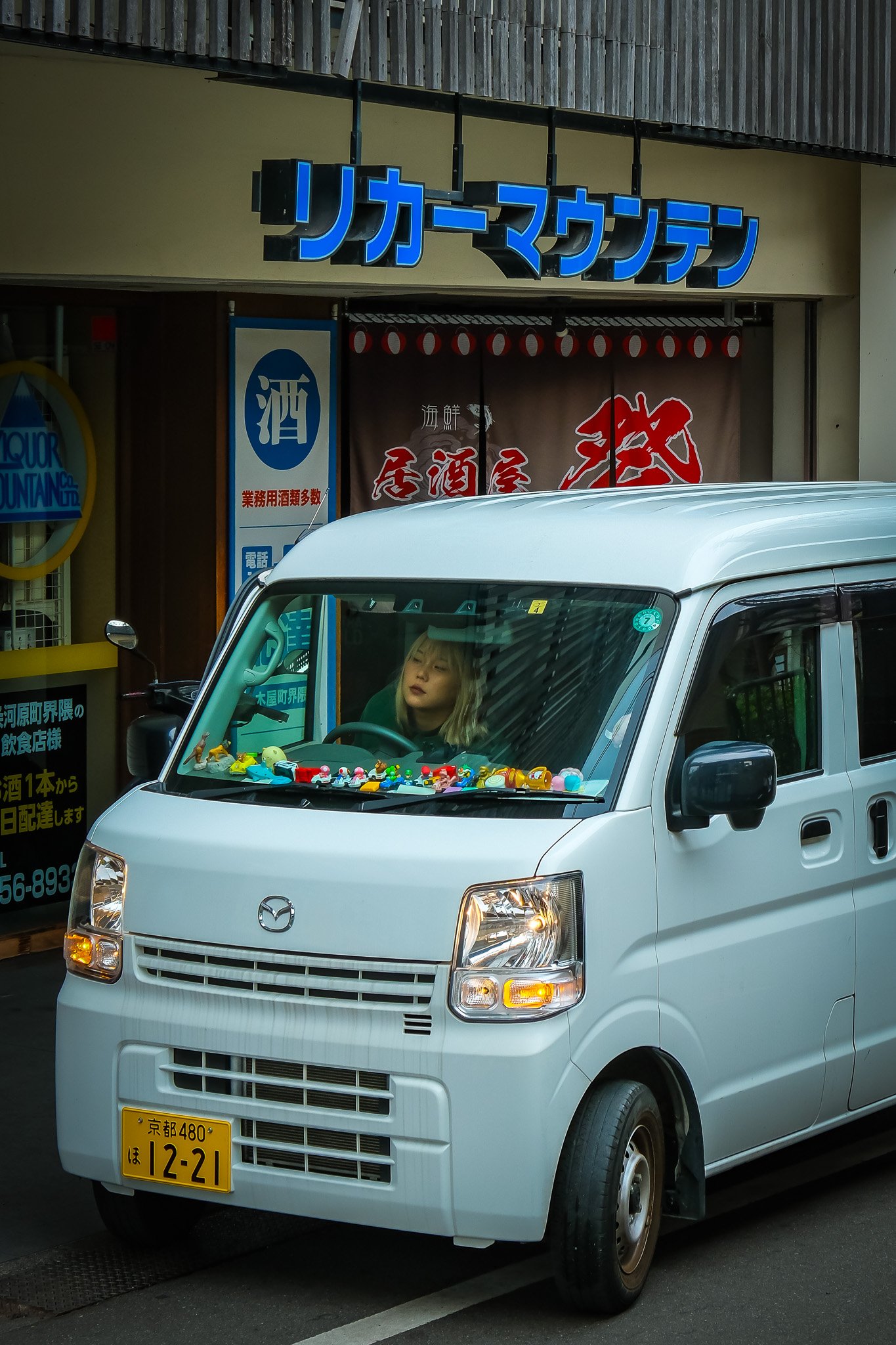 A white Mazda kei car parked on the street outside of Liquor Mountain storefront with toy figurines on the dashboard in Ginza Tokyo.  including one with the word '酒' (sake)