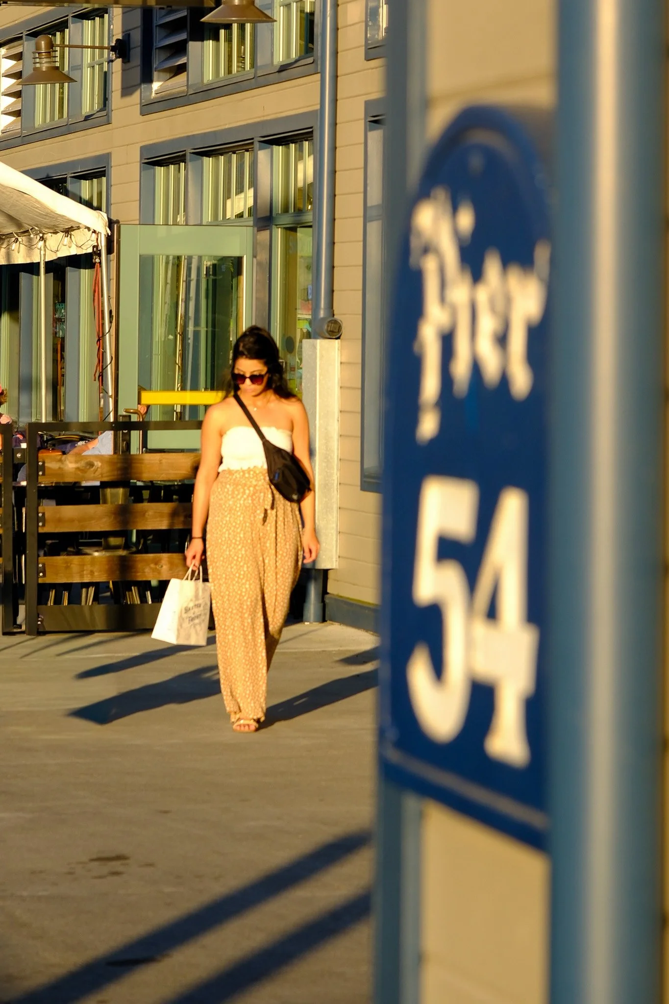 A woman with long dark hair, wearing sunglasses, a white strapless top, and the yellow patterned pants, walking on the sidewalk while carrying a shopping bag and a black shoulder bag in Seattle Washington along pier 54 in the summer 