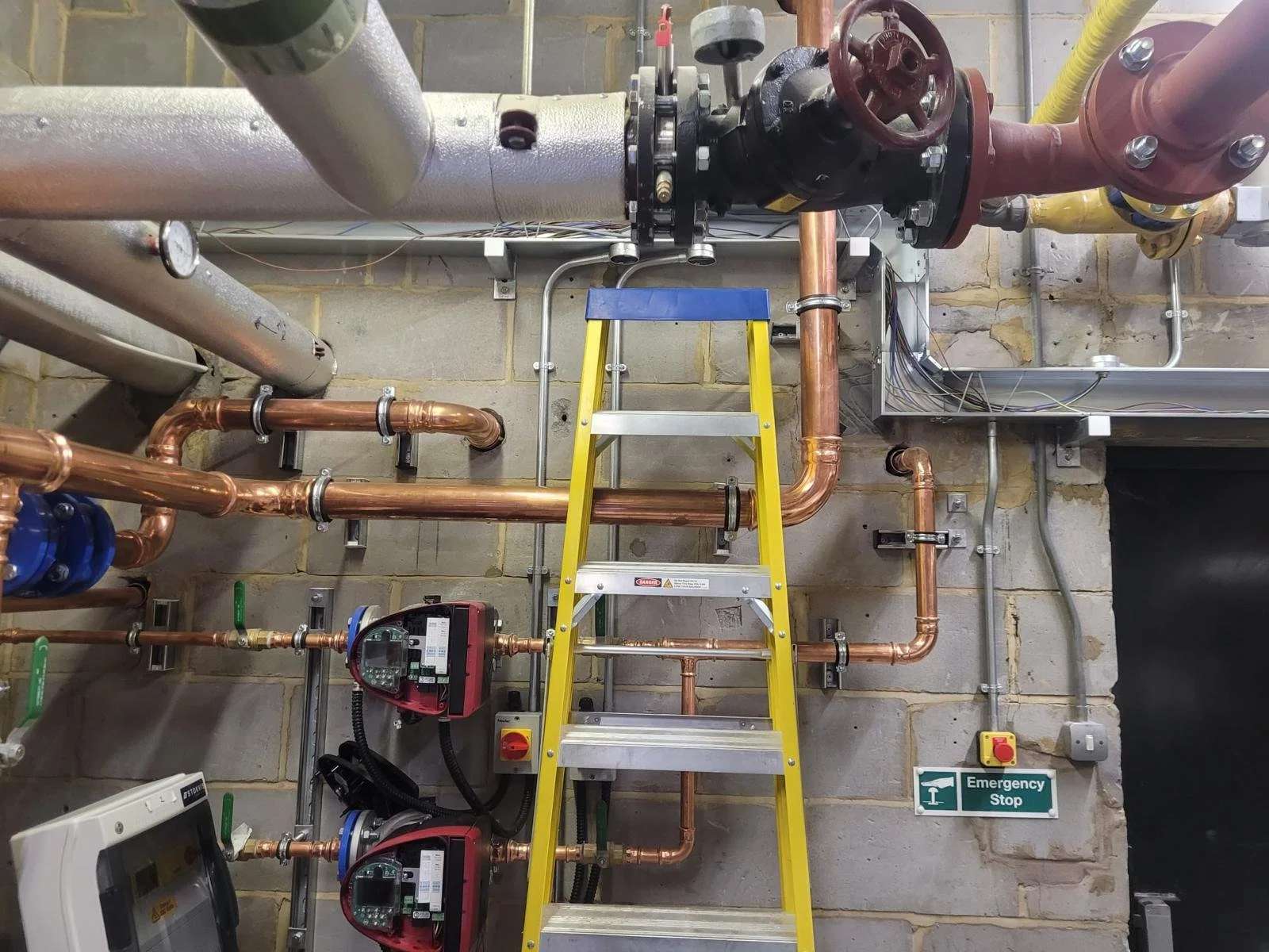 Industrial pipes and valves with a yellow stepladder and an emergency stop sign on a wall