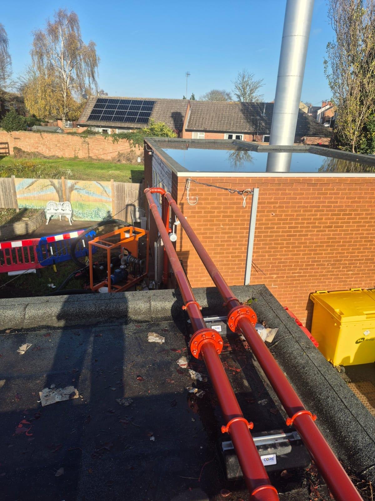 View from a rooftop showing red pipes connected to a building, with a brick wall, yellow equipment, and a neighborhood with solar panels on rooftops in the background.