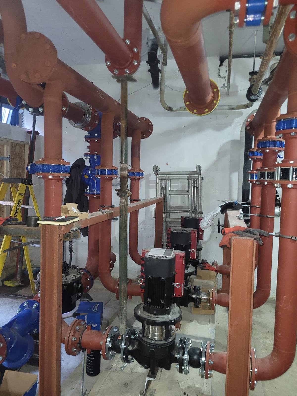 View of a plumbing system with large red pipes, valves, and a pump, under construction or installation in a building.