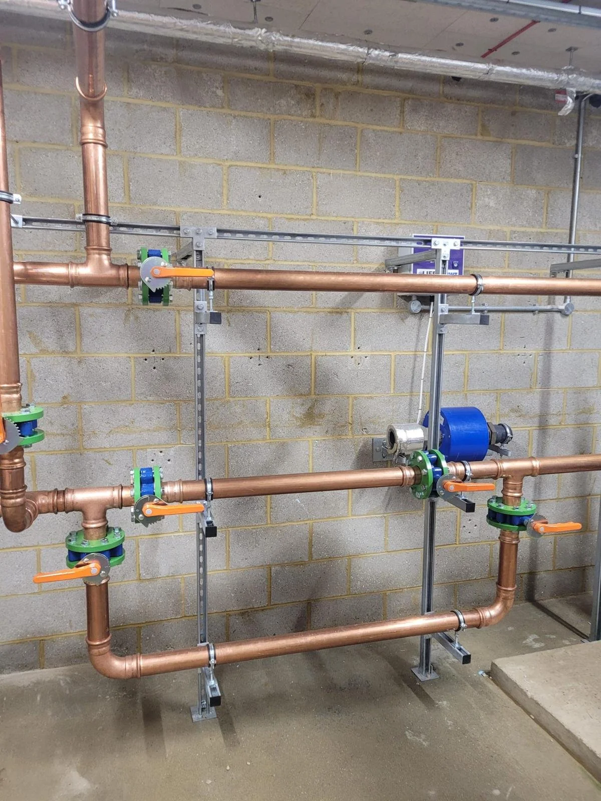 A plumbing setup with copper pipes, valves with orange handles, a blue pump, and metallic brackets mounted on a concrete floor against a brick wall.