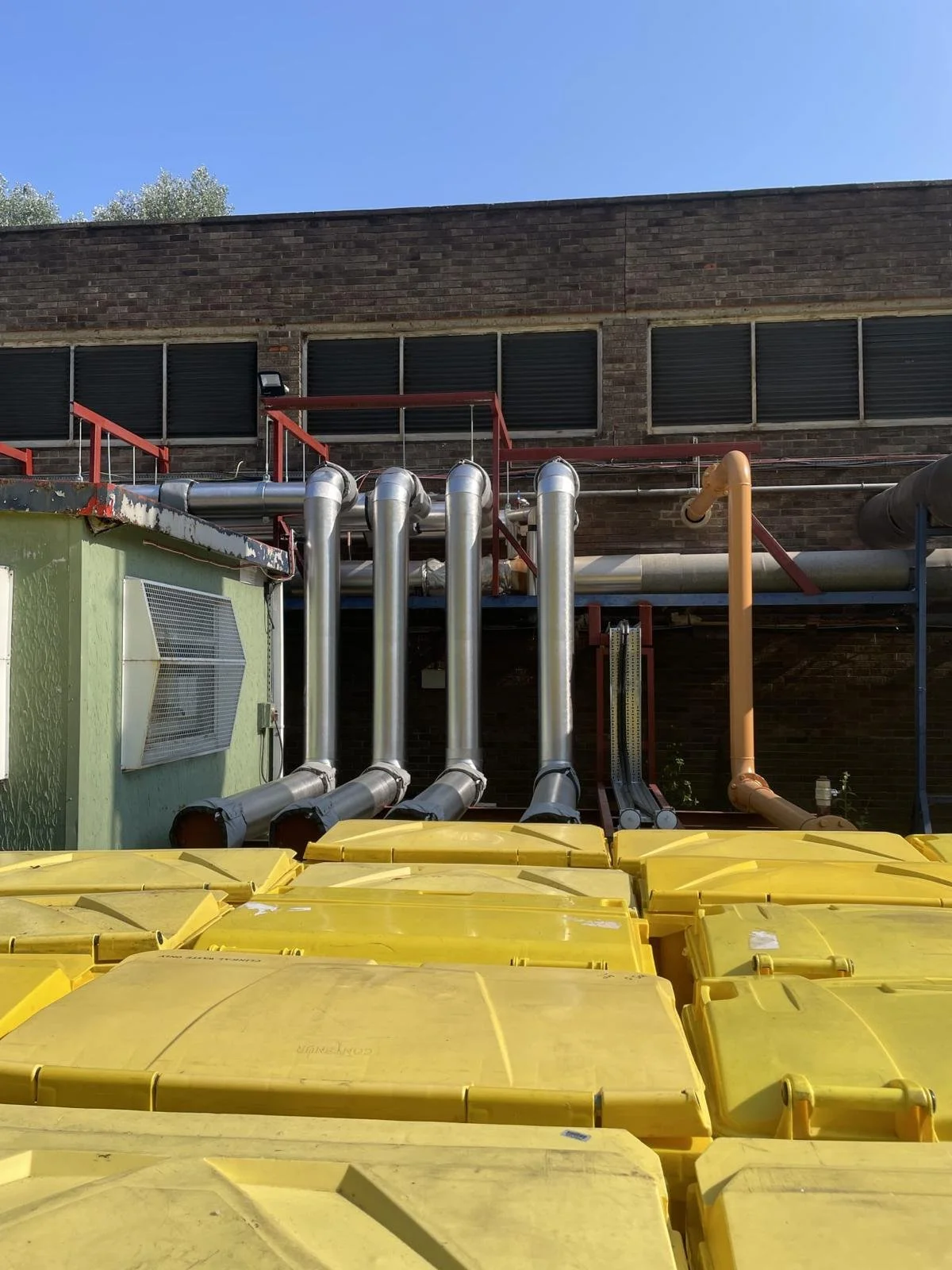 Industrial area with yellow storage bins in the foreground, metal pipes, a green building with a vent, and a brick building in the background, along with a clear blue sky.