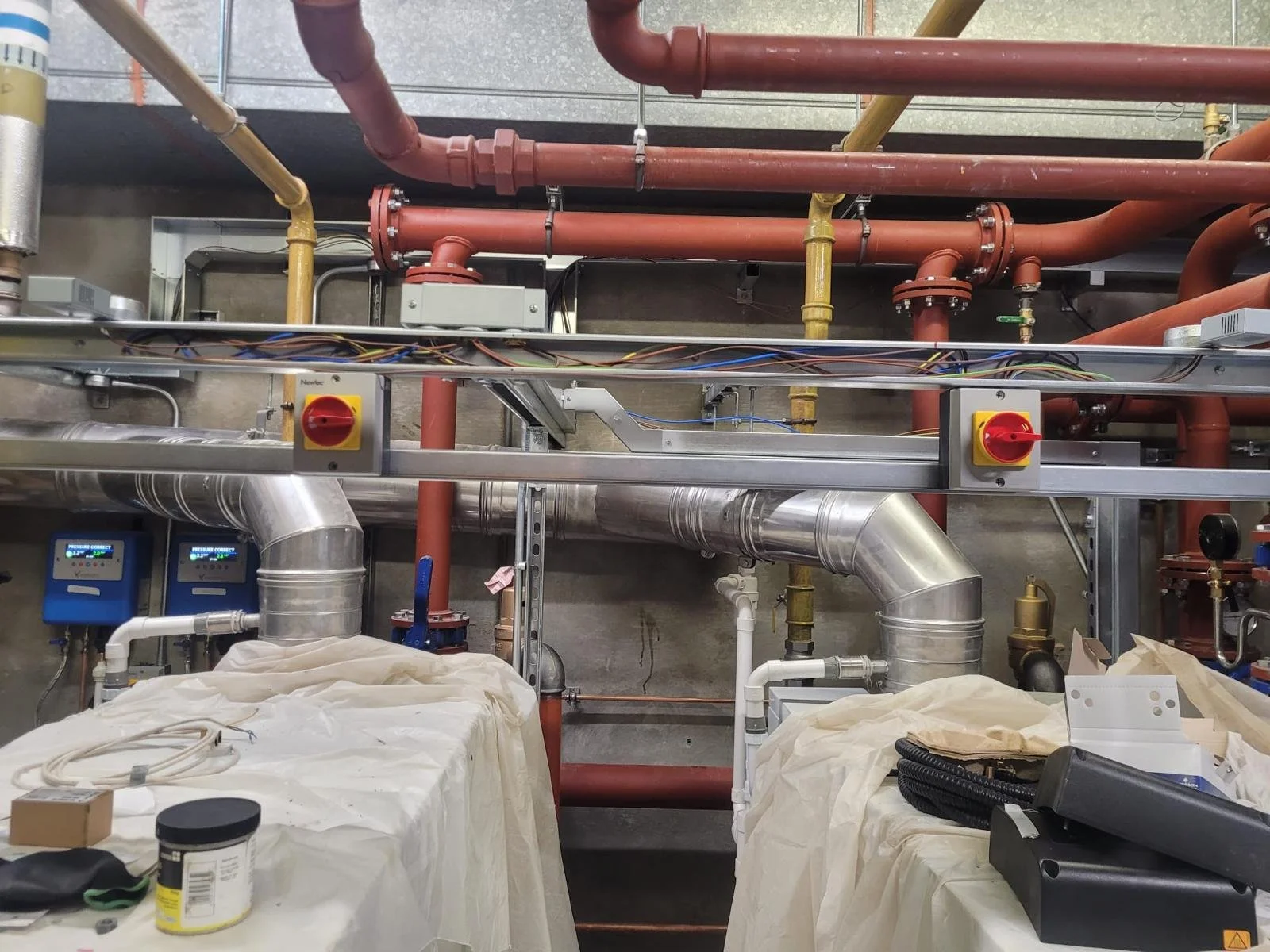 Industrial plumbing and piping system with red and yellow pipes, silver ducts, gauges, and switches in a mechanical or HVAC room.