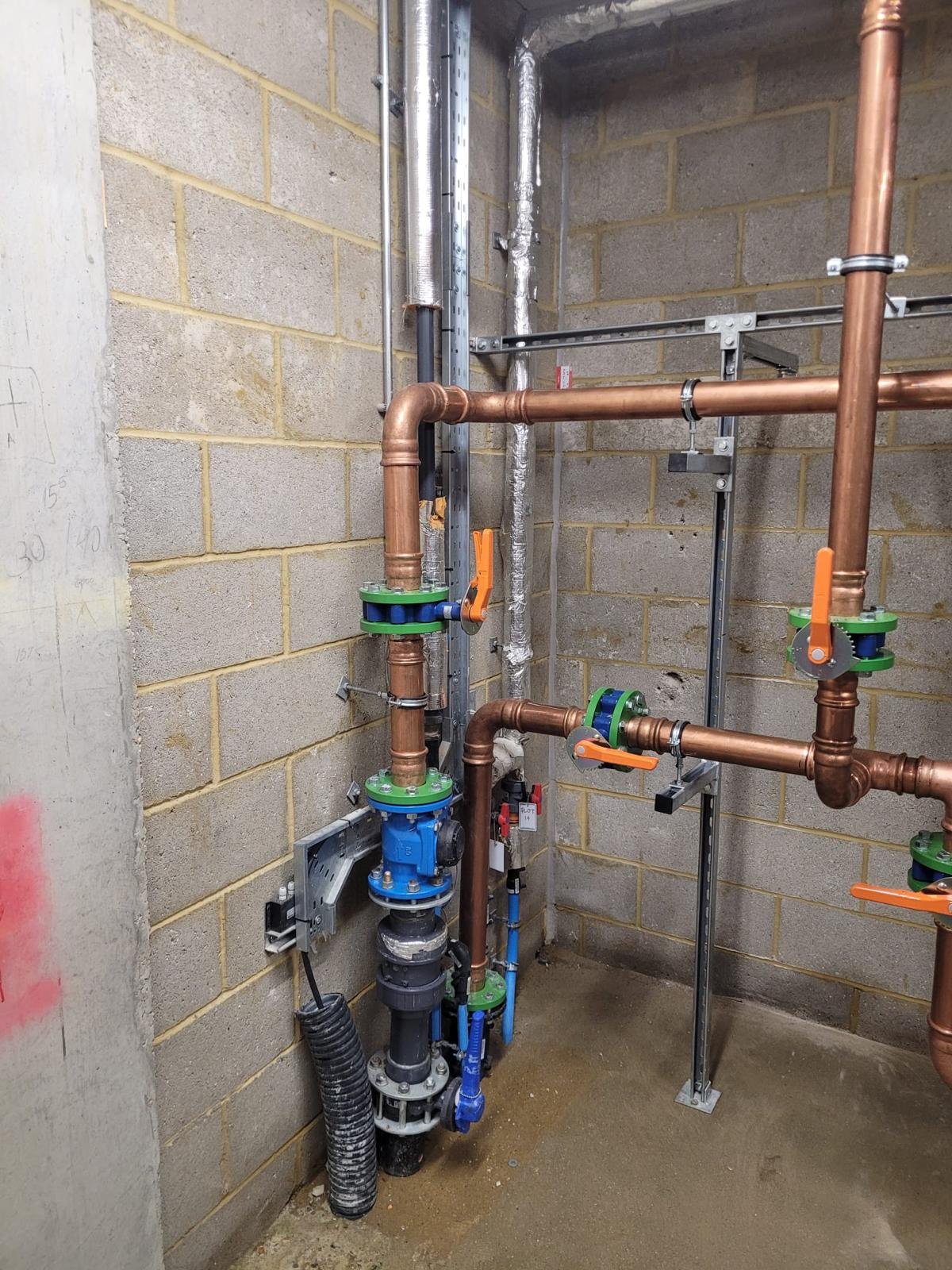 Indoor view of plumbing pipes and fixtures, including copper pipes, valves with orange handles, a blue valve, and various fittings and supports, against a cinder block wall.