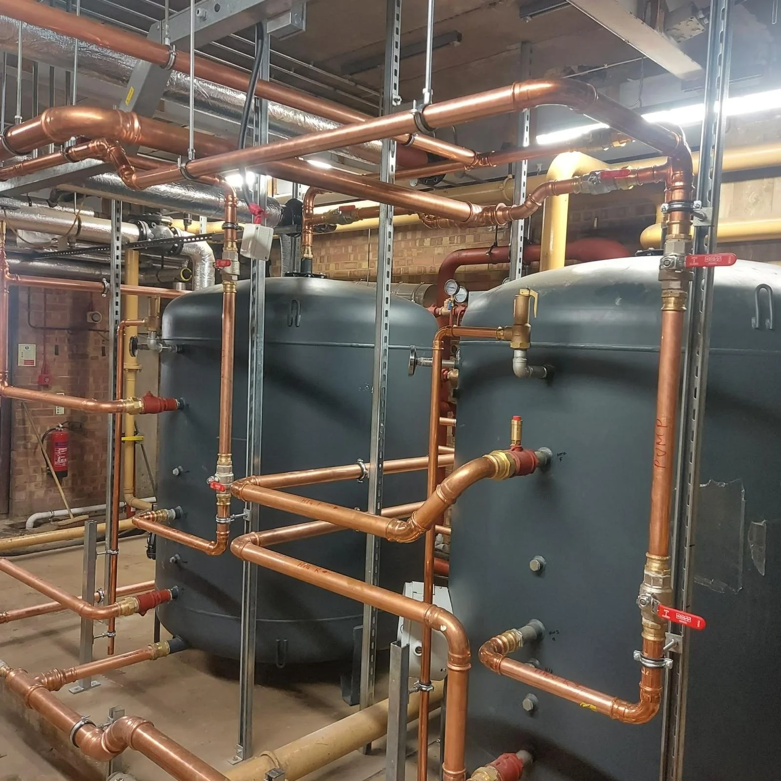 Interior view of a mechanical room with various copper pipes, large tanks, pressure gauges, valves, and metal framing supporting the piping system.