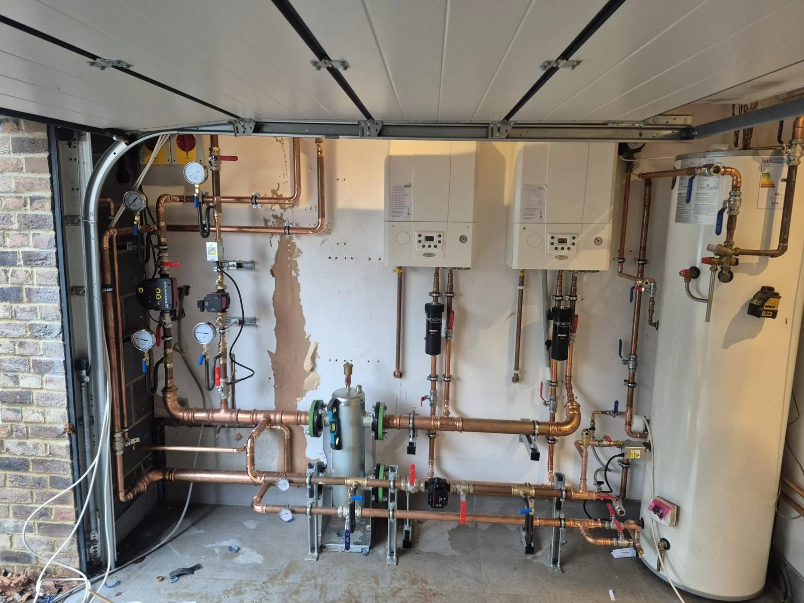 A plumbing setup with copper pipes, gauges, and meters, installed inside a room with unfinished wall and a ceiling with visible panels. There are two wall-mounted units and a large water heater on the right.