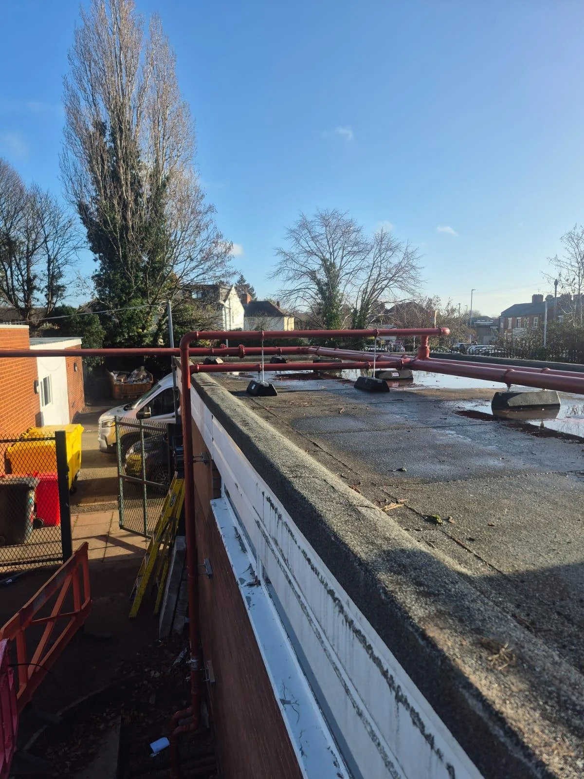 A rooftop with red pipes and black supports, surrounded by trees and houses in a clear, sunny sky.