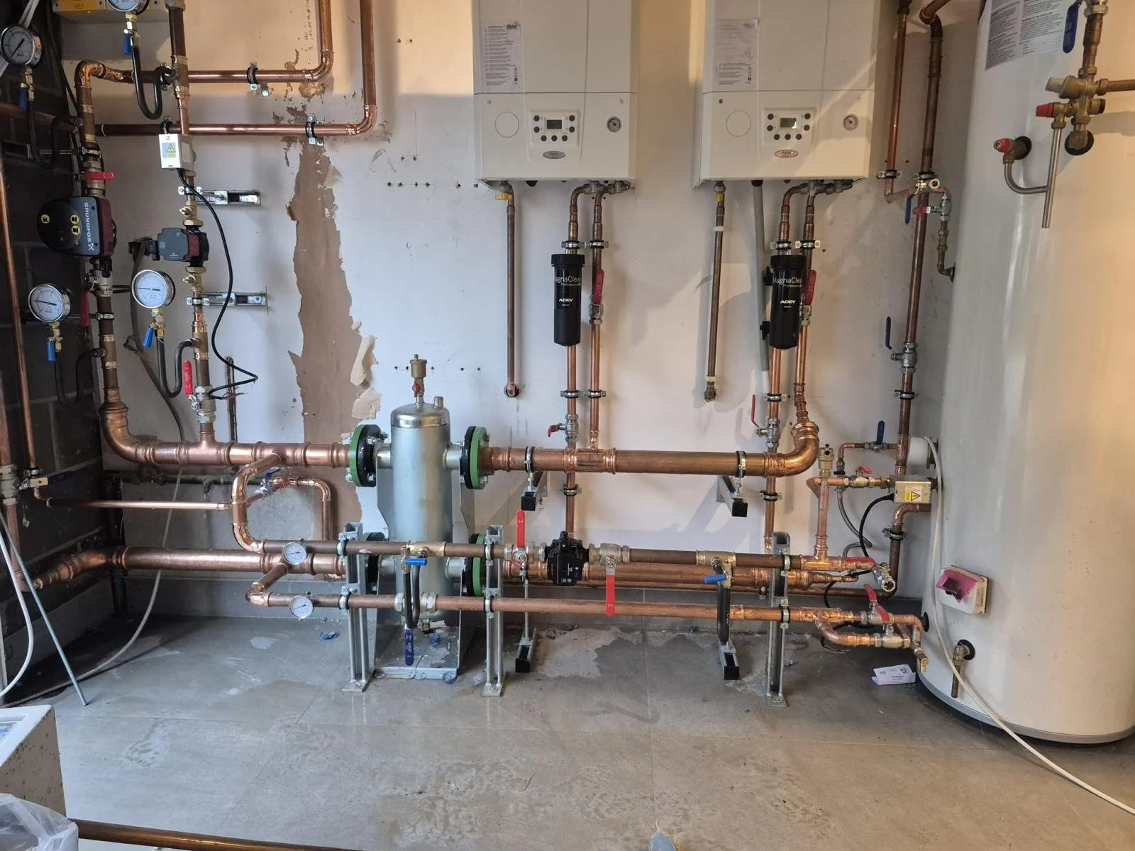 Plumbing and heating system with copper pipes, gauges, and two white wall-mounted boilers in a utility room.