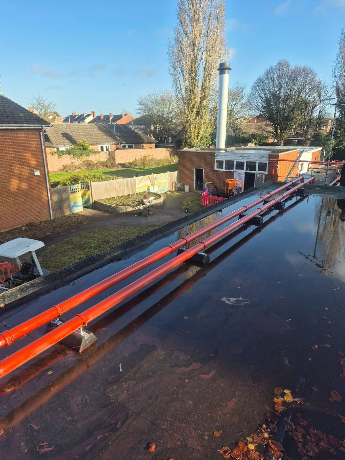 Rooftop view showing orange pipes and reflective water on the roof, with a backyard and buildings with chimneys and trees in the background under a blue sky.