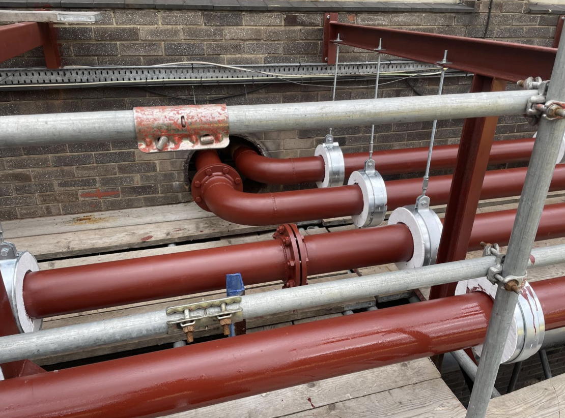 Construction scaffolding with red-painted pipes and metal clamps, attached to a brick wall with electrical wiring above.
